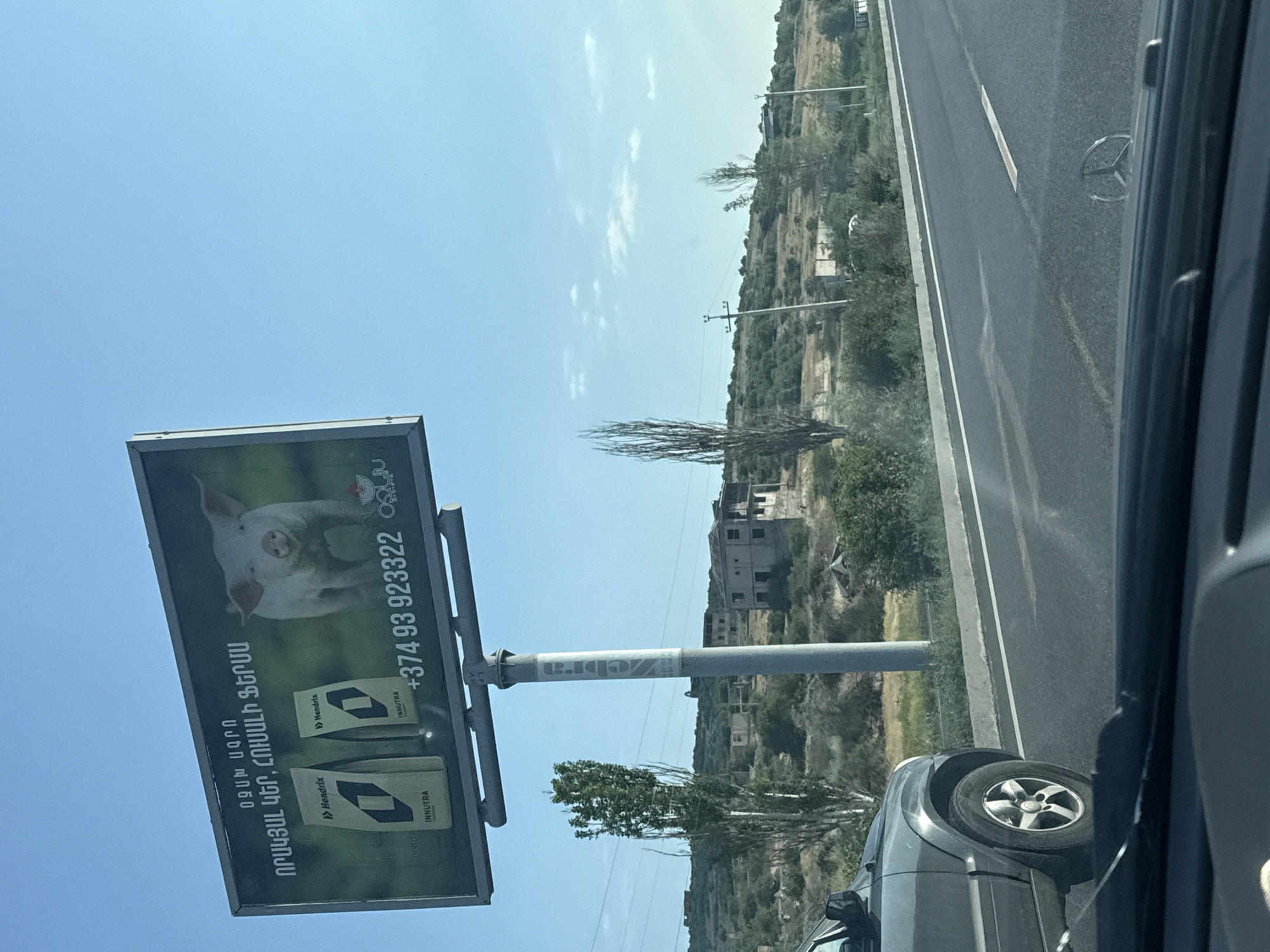 The image captures a billboard advertisement in Arzni, Armenia. The billboard features a close-up of a young pig against a dark green background. To the left of the pig are two bags of what appears to be animal feed, each branded with "Hendrix" and "INNUTRA." The text on the billboard is in Armenian. The billboard is mounted on a tall metal pole. In the foreground, the front of a dark gray SUV is visible, with its wheel turned slightly. The SUV is on a paved road, suggesting the image was taken from inside another vehicle. The background shows a hilly landscape with scattered buildings, sparse vegetation, and utility poles under a clear blue sky with a few scattered clouds.