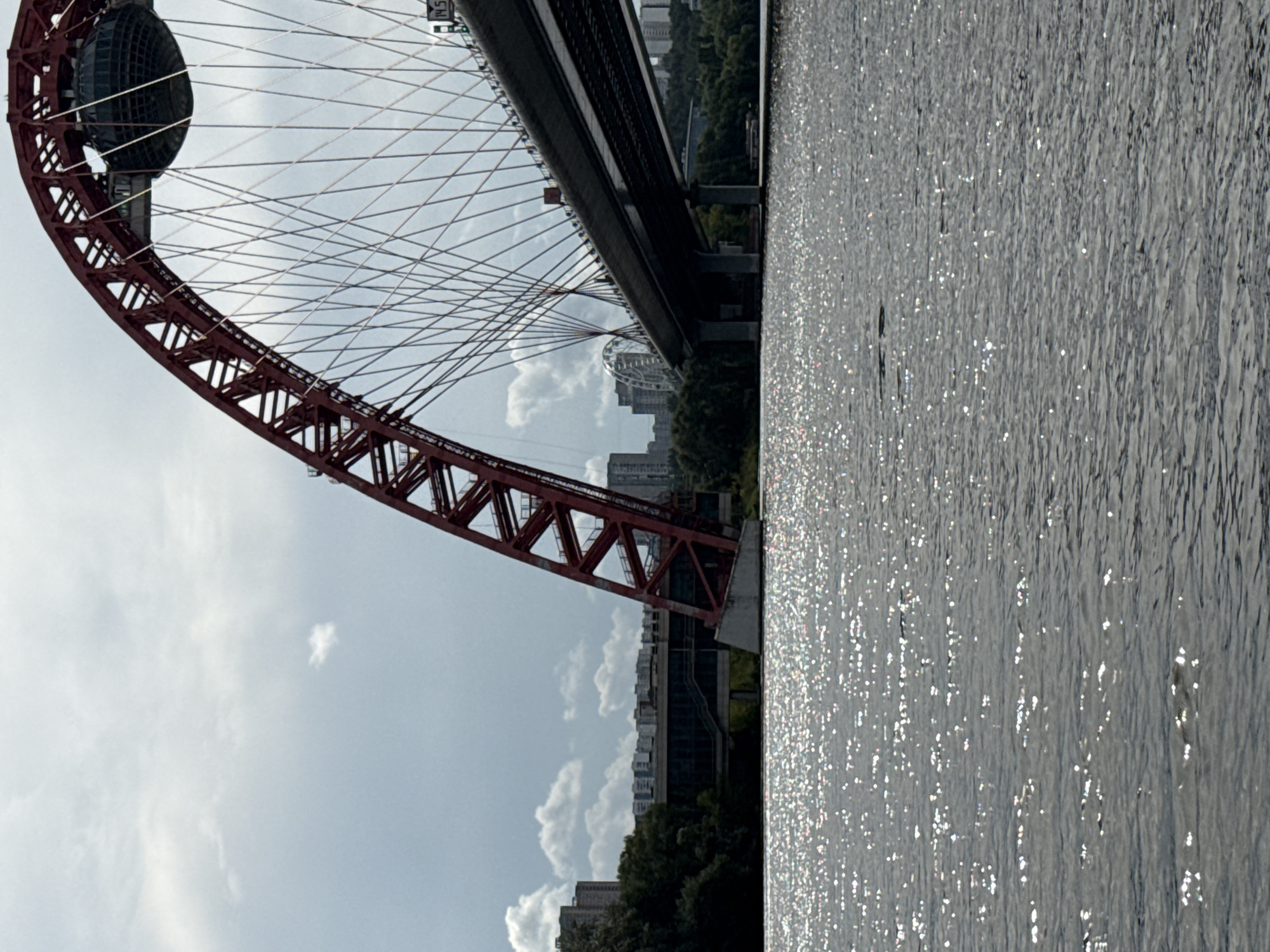 The image depicts the picturesque Zhivopisny Bridge in Moscow, Russia. The bridge, a prominent landmark, is characterized by its striking red arch and a spherical structure at its apex. The cable-stayed design of the bridge is clearly visible, with numerous cables extending downwards to support the roadway. In the background, a dense urban landscape of apartment buildings and trees lines the riverbank. The water in the foreground is shimmering, reflecting the sunlight and creating a dynamic texture. The sky is partly cloudy, with scattered white clouds against a pale blue backdrop. The overall scene captures the architectural grandeur of the bridge set against a natural and urban environment.