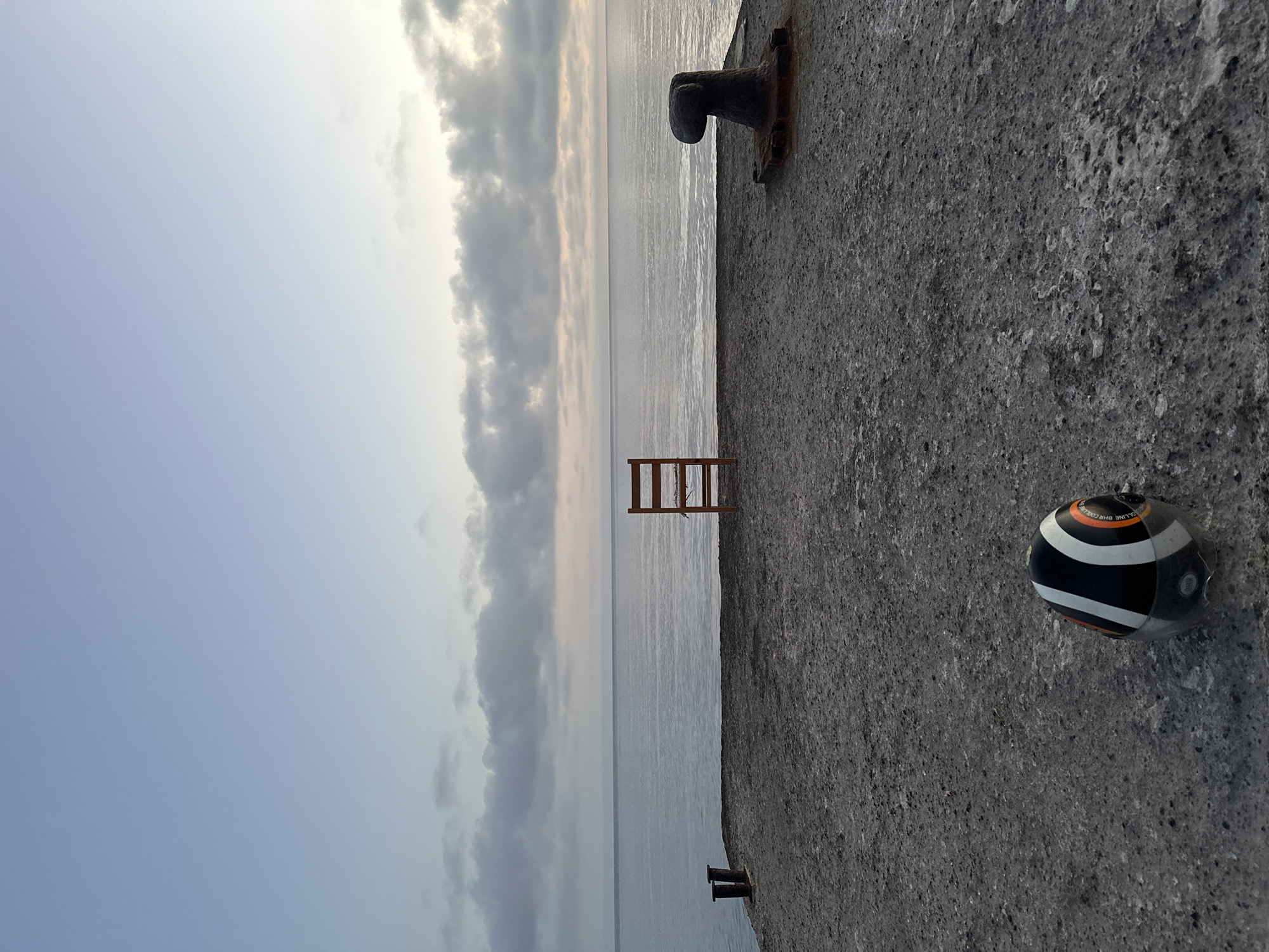 This media file captures a tranquil coastal scene, likely a pier or dock, at either dawn or dusk. The image is composed of three main horizontal sections: a concrete pier on the right, a calm body of water in the middle, and a partially cloudy sky on the left.
On the concrete pier, a rustic wooden chair is positioned facing the water, suggesting a spot for observation or rest. Further along the pier, a dark, weathered metal bollard, used for mooring boats, is prominently visible. In the foreground on the pier, a black and white striped nautical buoy or fender is resting, with partially visible text "COLLINE" and "BHB.CHROME" printed on its surface. Another smaller, dark metal fixture, possibly another bollard or mooring cleat, is barely visible further down the pier towards the horizon.
The water appears still and smooth, reflecting the muted light of the sky. The sky itself transitions from a clearer, lighter blue towards the left, to a cloudier, slightly warmer hue near the horizon, indicative of either sunrise or sunset. The clouds are soft and scattered, casting a gentle light over the scene.
The absence of people and the calm environment evoke a sense of peaceful solitude. The concrete texture of the pier is rough and worn, contrasting with the smooth water and soft sky. The overall impression is one of quiet contemplation by the sea.