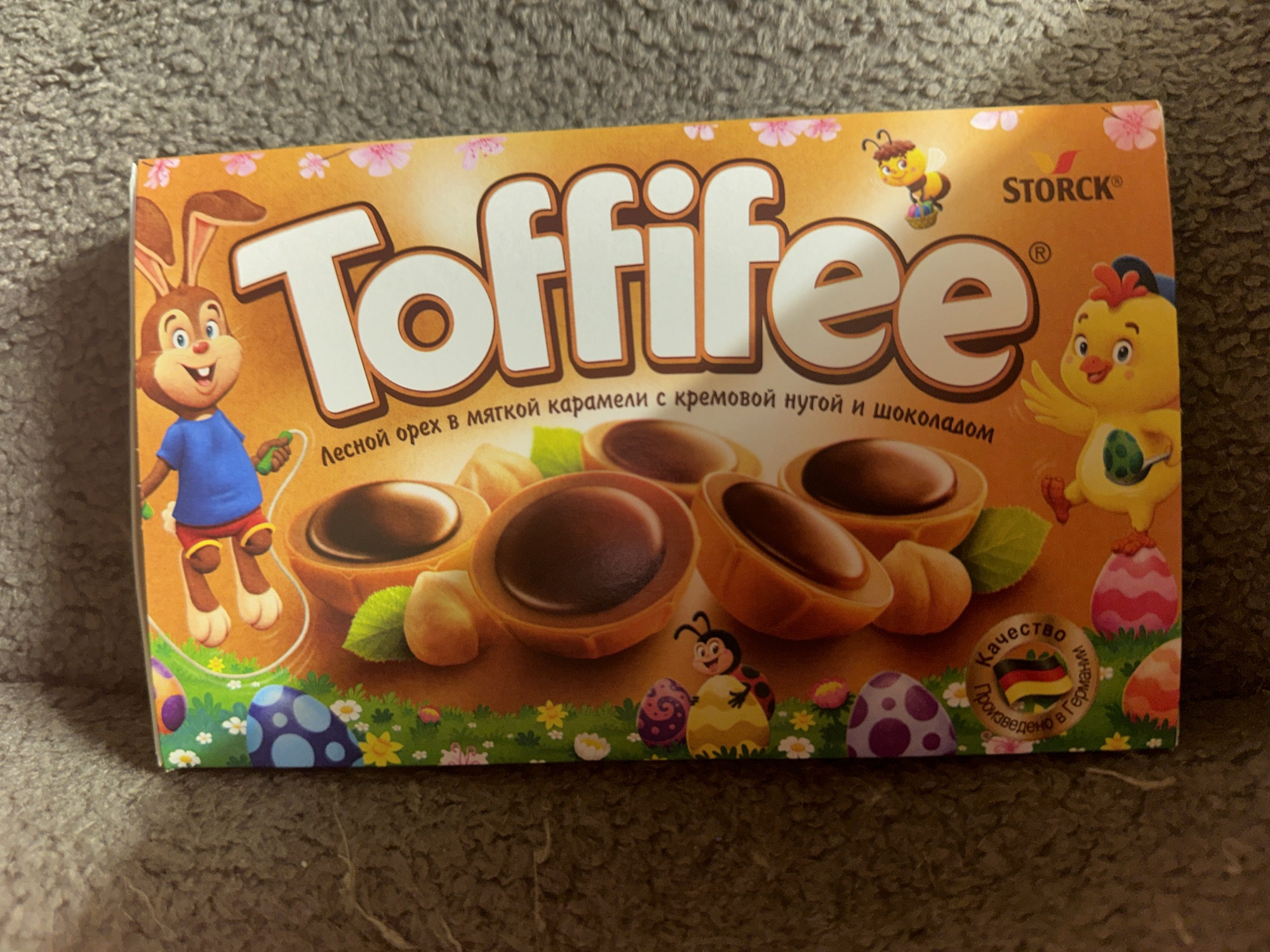 The image depicts a box of Toffifee candies, likely intended for an Easter promotion. The box features cartoon illustrations of a bunny, bee, and chick, along with images of Easter eggs. The prominent branding includes the "Toffifee" name and the Storck logo. There's a description of the product in Russian: "Лесной орех в мягкой карамели с кремовой нугой и шоколадом" (Hazelnut in soft caramel with creamy nougat and chocolate). The box includes a German flag indicating the product's origin. No information suggests the location of Moscow, Russia, nor the presence of individuals or specific environmental elements.