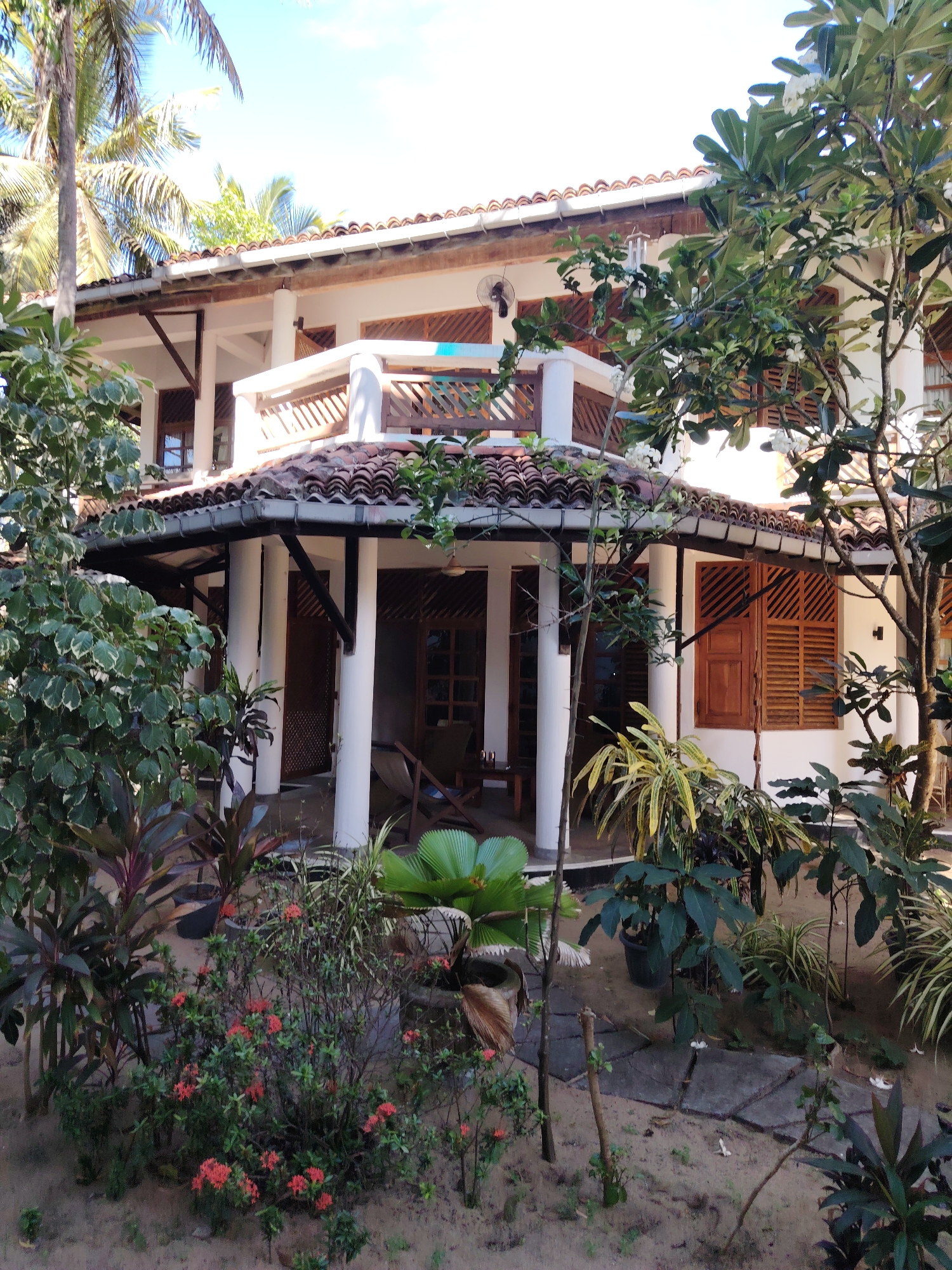 An outdoor, eye-level, medium shot captures a white, two-story building in a tropical environment. The building features red tile roofing, white pillars supporting a covered porch, and wooden louvered windows. Lush green foliage, including palm trees and flowering bushes, surrounds the structure. The ground is covered in sand and stepping stones, suggesting a garden or courtyard area. The sky is visible and appears bright blue, indicating daylight. There are no people or animals visible in the scene.