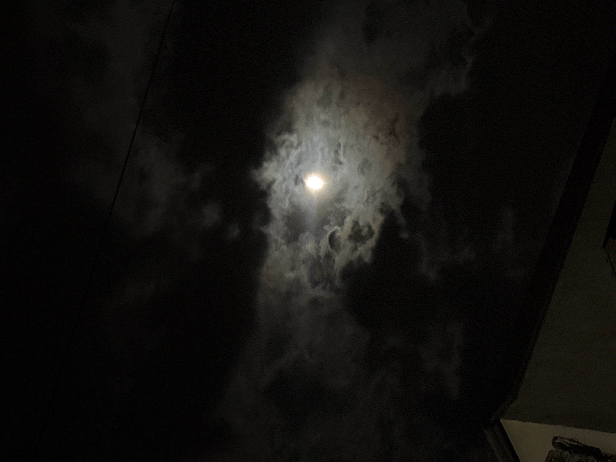 The image depicts a night scene with the moon obscured by clouds. The moon is visible as a bright, slightly diffused light source in the center. Dense, dark clouds surround the moon, creating a dramatic contrast. A faint, straight line is seen on the left, possibly a power line. The bottom right shows a glimpse of a dark, angular structure, suggesting a building or part of a building. The location is Unknown, Unknown.