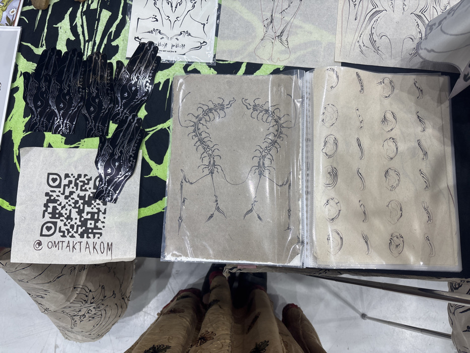 The image depicts an overhead view of a display table in Ban Rong Ruea Kham, Thailand, covered with a black fabric featuring a neon green abstract pattern. On the table, various merchandise is arranged. In the upper left quadrant, a square piece of light brown paper exhibits a black QR code above the text "@OMTAKTAKOM". Adjacent to this, multiple black, hand-shaped objects with intricate silver wavy patterns are stacked and spread. Below these, two clear plastic sleeves contain sheets of light brown paper. The top sleeved sheet displays a black line drawing of a segmented, centipede-like creature forming a circular or heart-shaped motif, flanked by spear-like appendages. The bottom sleeved sheet presents multiple rows of smaller, distinct black line drawings, including wavy lines and irregular shell-like or organic shapes. To the right, additional sheets of paper with black line art, some resembling skeletal or anatomical structures and abstract wave patterns, are visible. On the far left, the lower body of an individual is partially visible, wearing a patterned light-colored garment and red footwear. A thin, silver-colored metallic pole is visible in the lower left foreground, and a light-colored floor extends beneath the display.