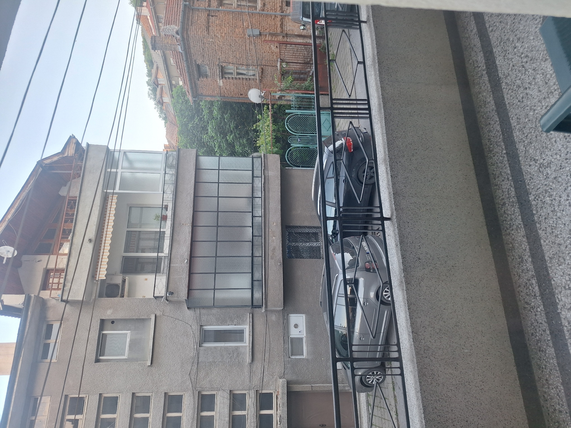 This is a view from a balcony in Asenovgrad, Bulgaria. The image depicts two cars parked in front of a multi-story building with a mix of modern and older architectural styles. There is a fence or railing in the foreground, indicating that the photo was taken from a higher vantage point. The buildings appear to be located in a residential area. There is green vegetation visible in the background. The weather appears overcast.