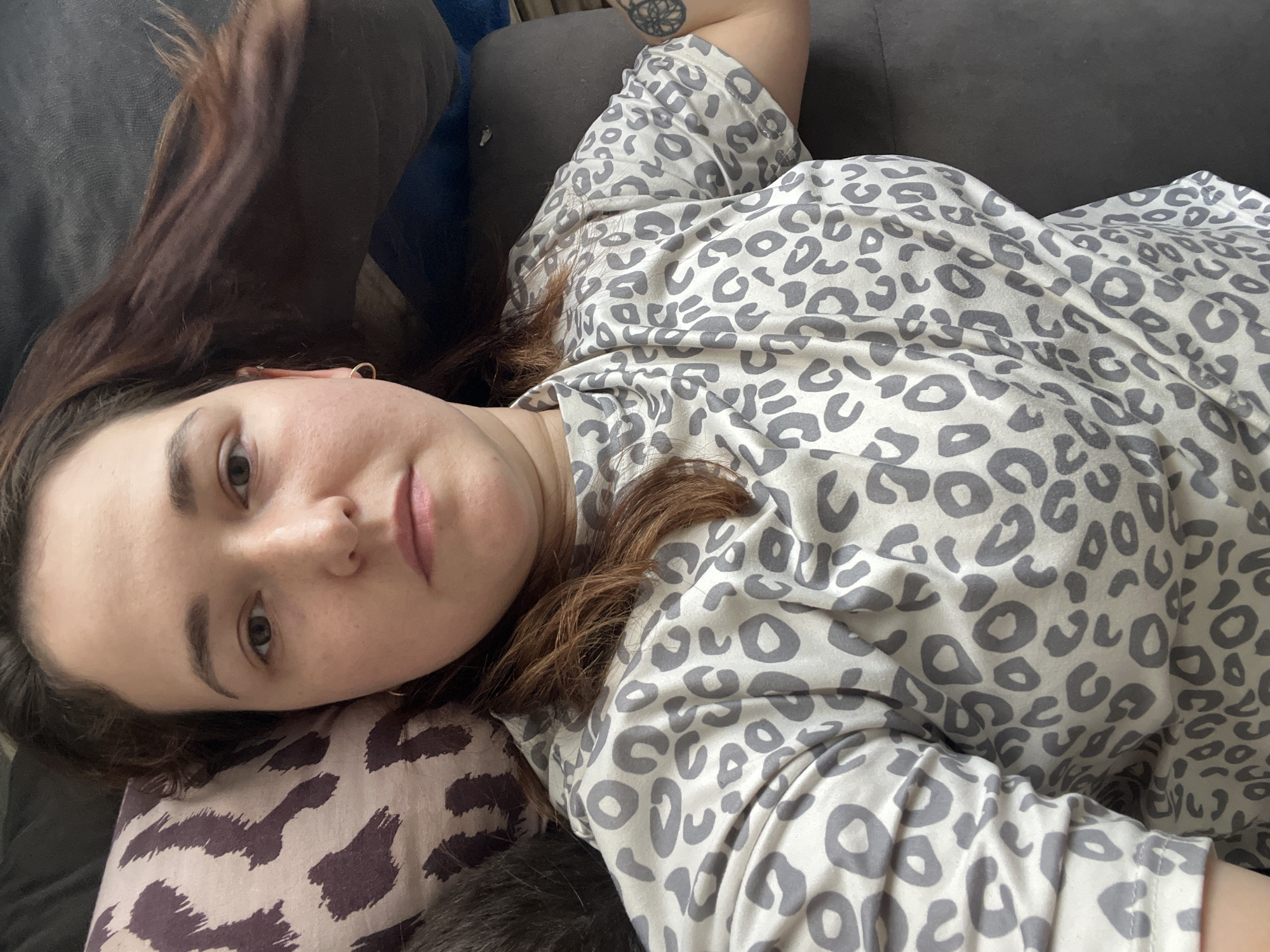 This is a close-up, overhead selfie of a young woman lying on a couch. She has long, dark brown hair that is spread out around her head. She is wearing a light-colored t-shirt with a gray leopard print pattern. Her head is tilted, and she is looking towards the camera. Her expression is neutral.
The woman is lying on a dark gray couch. To her left, there is a pillow with a dark brown and cream-colored leopard print pattern, which is partially obscured by her head. Her right arm is extended, and a portion of her left arm is also visible. A tattoo with a circular design is visible on her upper right arm.
The lighting suggests it could be daytime, possibly indoors as there are no clear indications of natural light or weather. The overall scene depicts a casual, relaxed moment, likely at home. The location is identified as Chelyabinsk, Russia. There is no visible text in the image.
