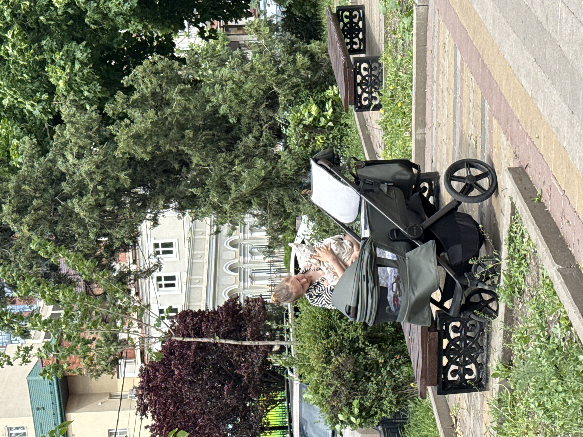 A woman in a zebra-print top is bent over a stroller next to a park bench. The stroller is dark green and black, and its canopy is partially open. The bench has decorative black cast iron legs. The scene takes place on a paved walkway in a park or garden, with trees and bushes visible in the background. A building with white arches is visible behind the trees, suggesting the location is within the city of Rostov-on-Don, Russia.