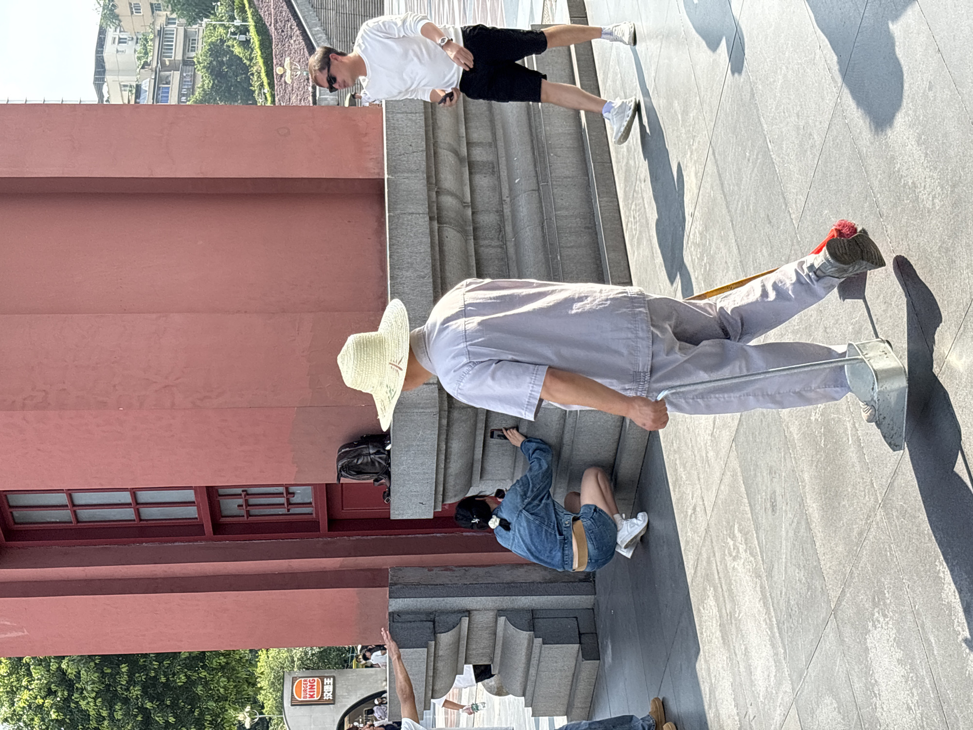 In Yuzhong District, China, a person wearing a straw hat and a light-colored uniform, holding a dustpan and broom, walks away from the camera on a paved walkway. To the left, a woman in a denim jacket and shorts kneels and takes a picture with her phone. Behind her, a black backpack rests on a stone ledge. To the right, a man in a white long-sleeved shirt and black shorts, wearing sunglasses, walks away from the camera. In the background, a large red building with windows is visible, and further back, buildings and trees on a hillside are present. A Burger King sign is visible on the left side of the image.