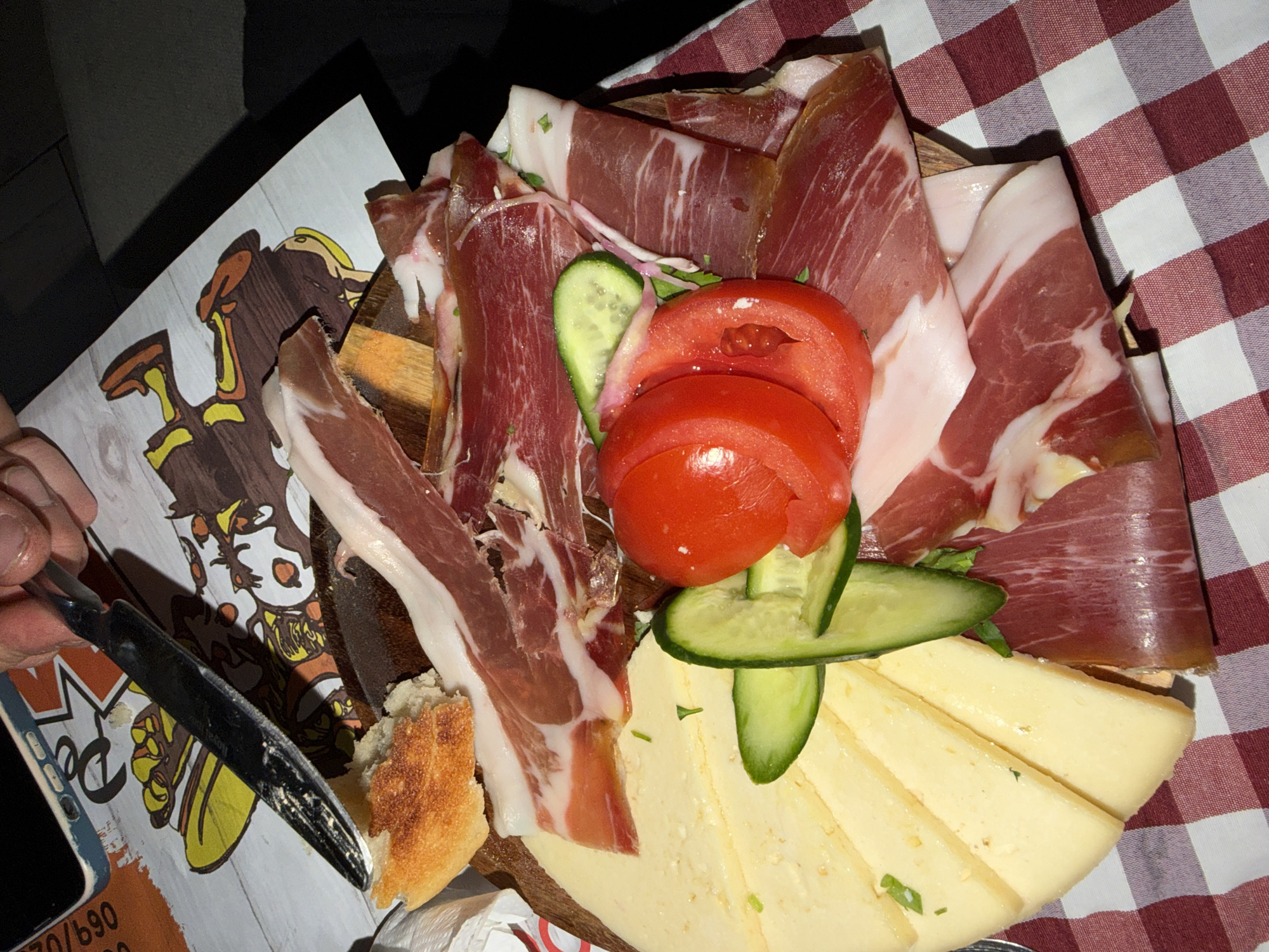 A wooden board of food is presented indoors on a red and white checkered tablecloth. The board is piled with slices of ham, tomato slices, cucumber slices, and wedges of cheese. A knife lies to the left of the board, near a piece of bread and a sheet of paper with a cartoon graphic. The setting is likely a restaurant or dining establishment, and based on information not explicitly visible in the image, this scene is in Budva, Montenegro. A hand is visible holding the knife.