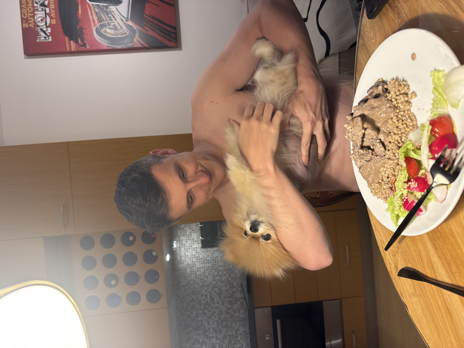 A man holding a small, fluffy, light brown dog, smiles at the camera. He's shirtless and standing in a kitchen with light wood cabinets. A plate of food is on the table, consisting of buckwheat, meat, and a salad with tomatoes and radishes. A poster of a car race hangs on the wall. The kitchen has recessed lighting and a backsplash of small, gray tiles.