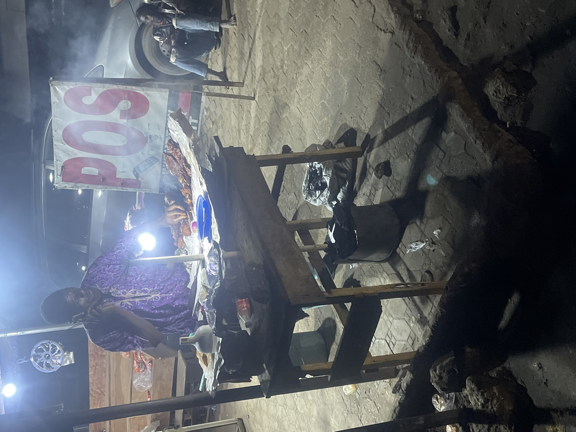 The image depicts a nighttime scene in Abraka, Nigeria, featuring a street vendor operating a roadside grill. The vendor, a man wearing a purple patterned shirt, is seen on the left side of the frame, seemingly on a phone call. He stands behind a makeshift wooden table displaying grilled meat. A POS (Point of Sale) sign is visible above the table. A white car is partially visible in the background, and a second person can be seen near the car. Debris and litter are scattered around the base of the grill stand and on the pavement. The street is dimly lit, with shadows cast by the grill and the table. The ground is paved with interlocking concrete blocks.