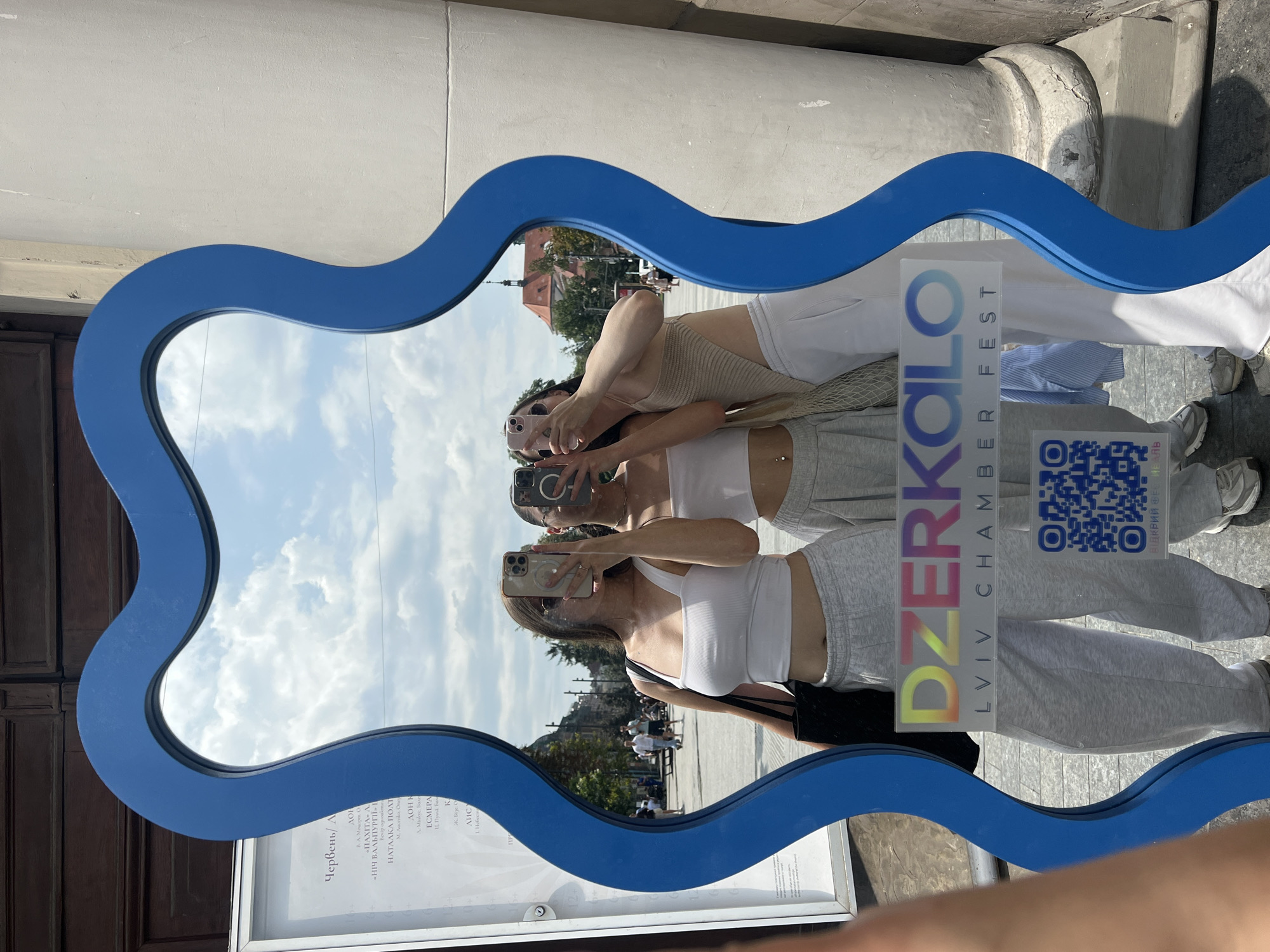 In Lviv, Ukraine, a wavy blue mirror reflects three young women taking selfies outdoors on a sunny day. The mirror is placed in front of a building with a large stone column on the right and a wooden door on the left. A poster with Ukrainian text is visible on the left side of the frame. The women are dressed casually in crop tops and sweatpants. The mirror has a sticker on it with the text "DZerkalo Lviv Chamber Fest" and a QR code. The background shows a street with people walking and buildings, indicating an urban setting.