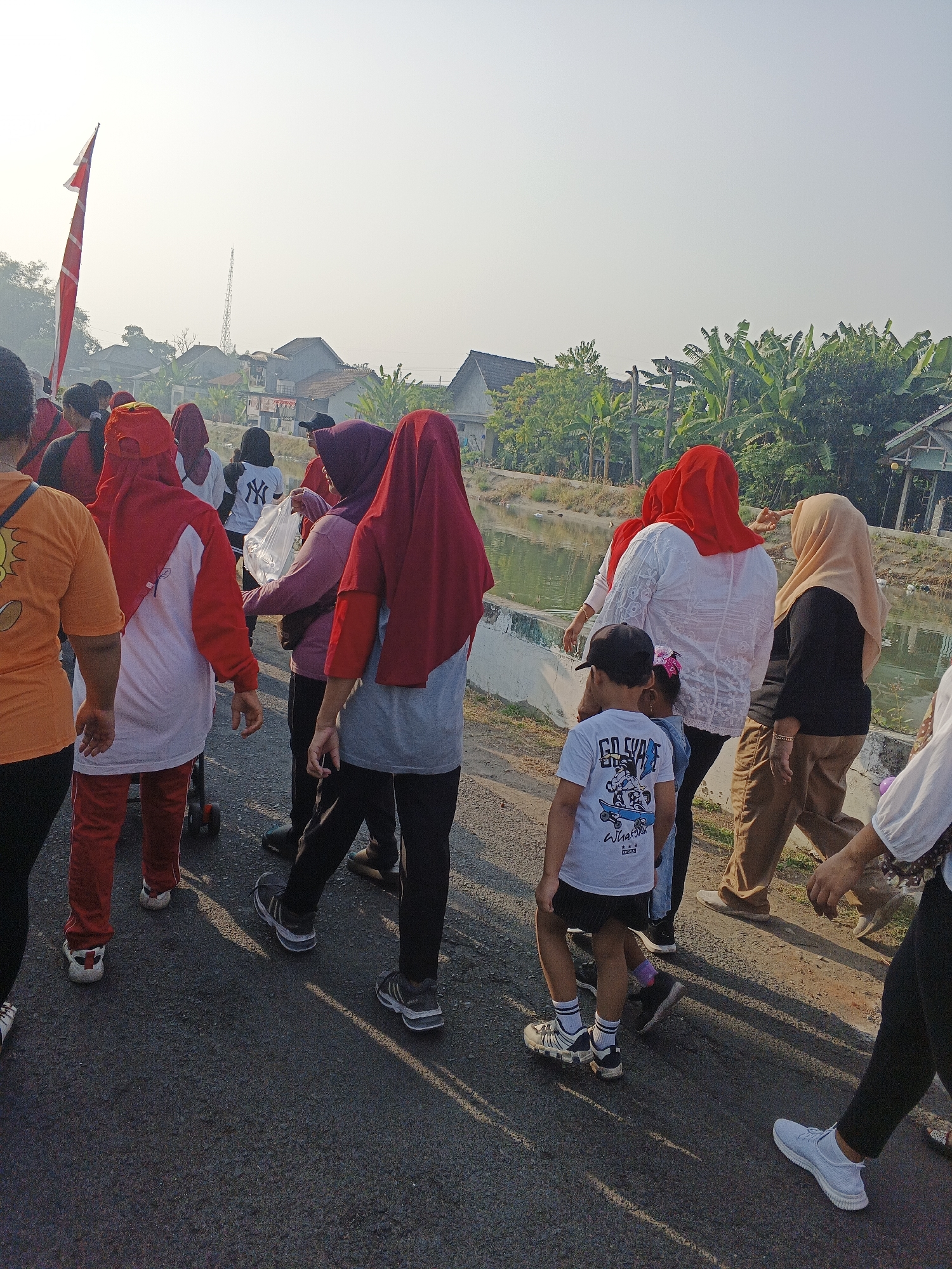 In Krian, Indonesia, a group of people are walking along a paved road next to a body of water. The group consists of adults and children, many of whom are wearing red and white or red headscarves, indicating a possible community event or exercise group. The people are dressed in casual athletic wear, suggesting they are participating in a morning walk or gathering. Some individuals are carrying bags, and one person is holding a white plastic bag. The environment shows residential buildings and lush green trees, including banana trees, lining the background, with a calm body of water to the right of the road. A red and white flag is visible on the left side of the image, adding to the festive or organized nature of the event. The sun is shining, casting long shadows on the road.