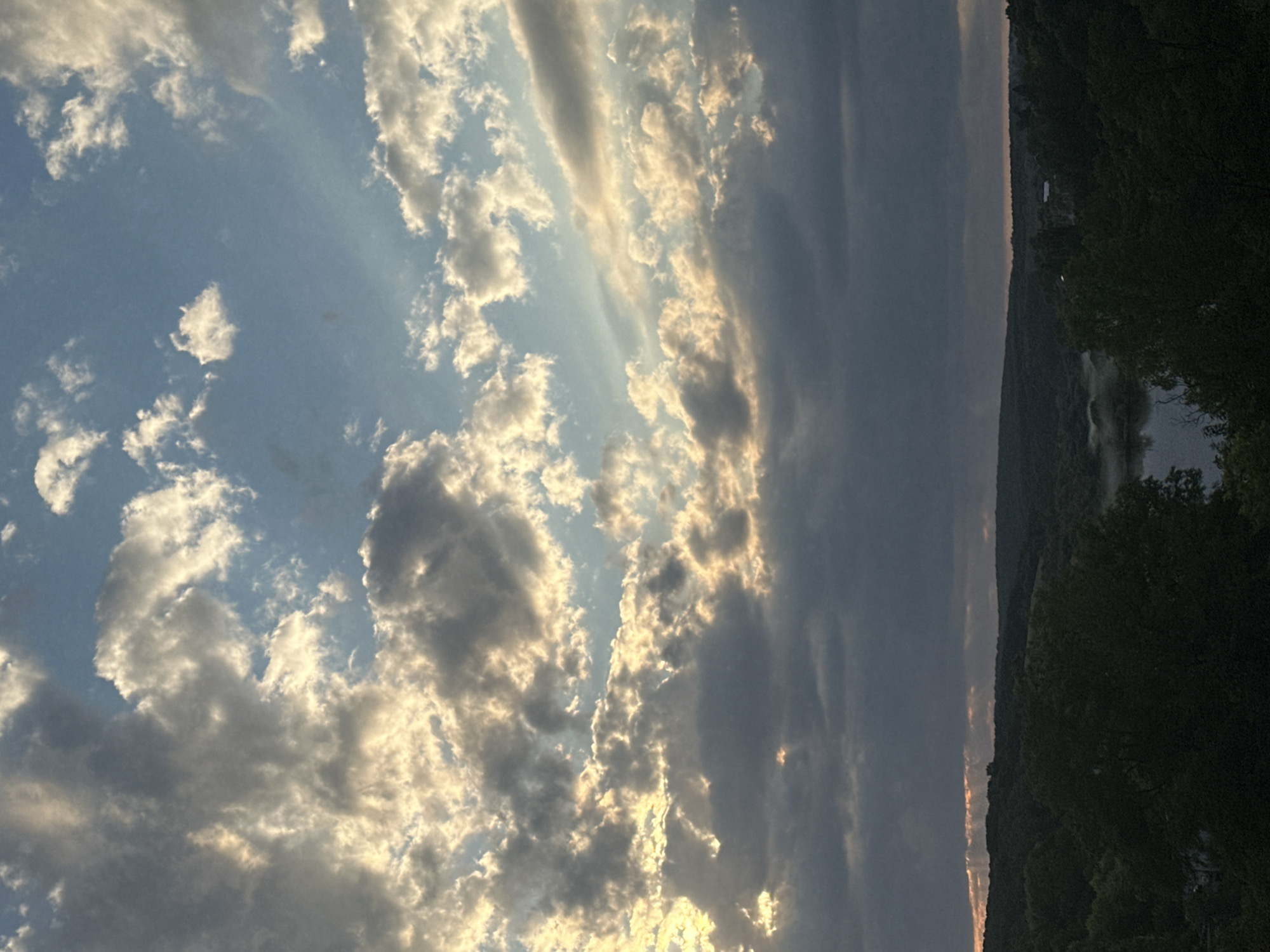 The image captures a panoramic view of the sky over Makiv, Ukraine, characterized by a dramatic interplay of light and shadow among the clouds. The upper portion of the image is dominated by a blue sky, punctuated by scattered white cumulus clouds and larger formations of dark, heavy clouds. Sunlight filters through some of the clouds, creating luminous, ethereal rays and highlighting the edges of others with a golden hue.
Below the sky, a dense, dark green expanse of trees stretches across the horizon, indicating a heavily wooded area. The terrain appears to be hilly or undulating. A body of water, possibly a river or lake, is visible in the middle ground, partially obscured by mist or fog, creating a soft, atmospheric effect. The water reflects the muted light of the sky. The overall impression is one of a natural landscape, likely a rural or semi-rural setting, observed during the late afternoon or early evening, given the quality of the light and the dramatic cloud formations. There are no people, specific structures, or discernible actions depicted in the image.