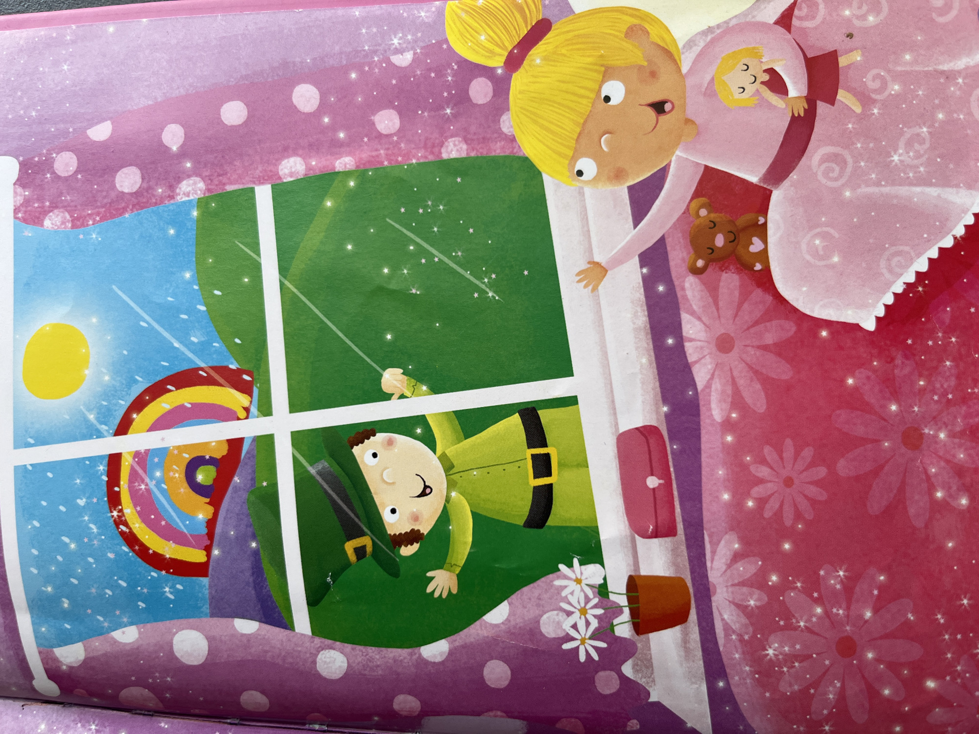 This media file is a close-up, angled shot of an illustration from a children's book. The artwork features a young blonde girl with a high ponytail, wearing a pink nightgown, peeking out from the top right of the frame. She is holding a doll with blonde hair and a red dress in her arms, and a small brown teddy bear with a heart detail is resting on her nightgown.
In the lower left quadrant of the image, through a windowpane, a scene unfolds. A smiling man dressed as a leprechaun, complete with a green hat, green tunic, and black belt with a gold buckle, is visible. He appears to be standing outside in a grassy area. Outside the window, a bright yellow sun shines in a blue sky dotted with white stars and sparkles, and a vibrant rainbow arcs across the sky.
The foreground of the illustration, at the bottom of the image, depicts a pink and purple patterned fabric with white polka dots, possibly representing a bed or blanket, with small white flowers in an orange pot. A pink pouch or purse is attached to the white frame of the window. The overall style is whimsical and colorful, with a strong sense of magic conveyed through the sparkles and starry elements. The illustration suggests a fantastical or dreamlike setting. There is no visible text.