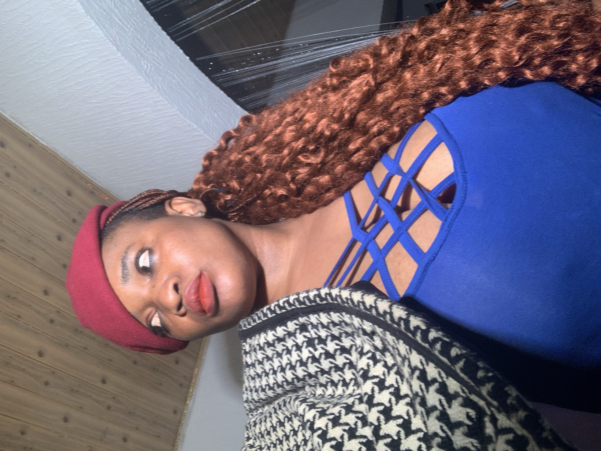 The image shows a woman posing indoors, rotated 90 degrees counter-clockwise. She has dark skin and is wearing a red beret. Her long, reddish-brown, curly hair, possibly extensions, cascades over her right shoulder. She is adorned with red lipstick and an earring on her visible ear. Her attire includes a royal blue top or dress featuring a distinctive criss-cross, strappy design on the shoulder and chest area. Partially visible, draped over her front, is a garment with a black and white houndstooth-like pattern.
The background features a light-colored wooden paneling on one side and a white, textured ceiling or wall on the other. Above her head and hair, a dark area with reflective or transparent material, possibly plastic sheeting or a window with reflections, is visible. The woman is looking upwards and slightly to her left, with a neutral to slightly alluring expression, suggesting she is posing for the camera. There is no visible text in the image. The lighting indicates an indoor setting, likely under artificial light.