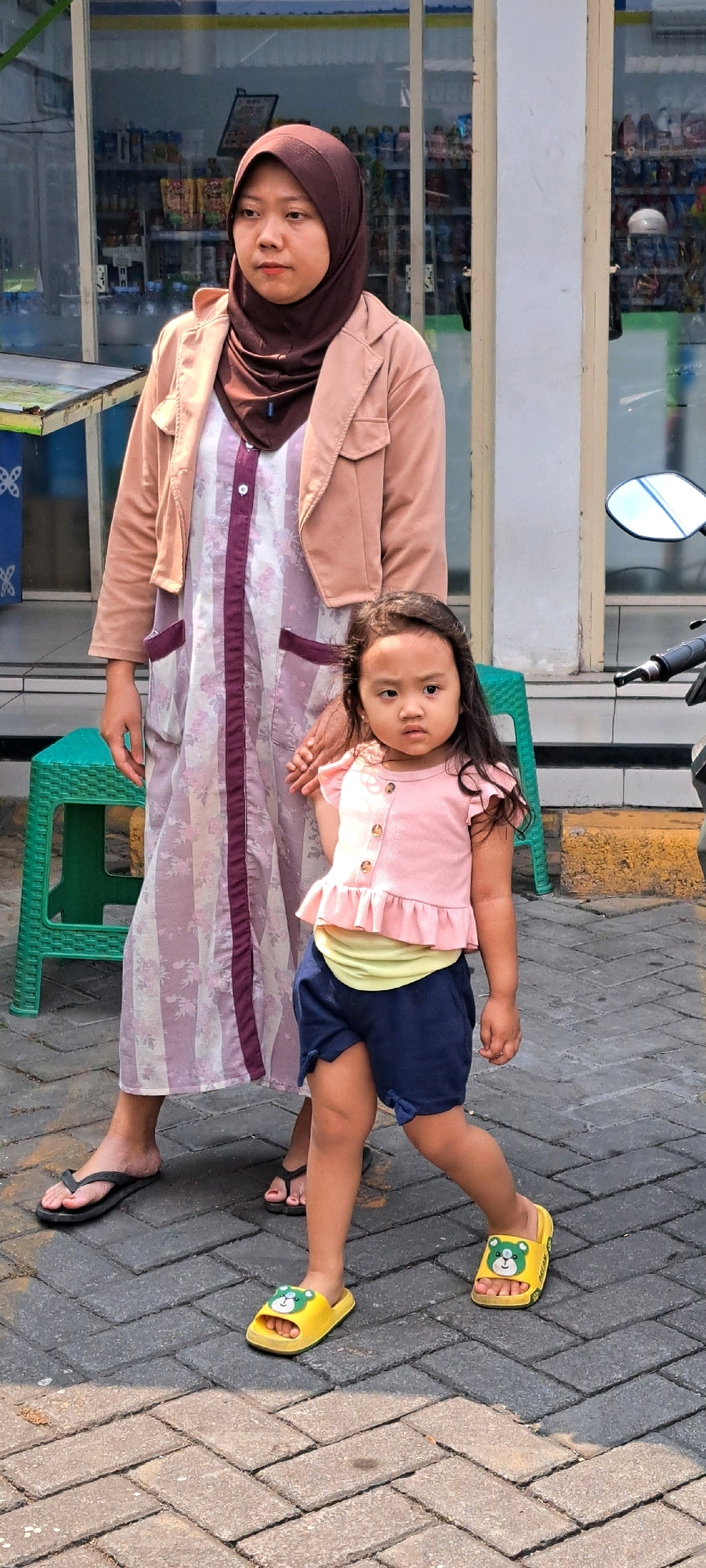 A woman wearing a brown hijab, a light pink jacket, and a long floral dress stands to the left, holding the hand of a young girl. The girl, facing forward with a neutral expression, wears a pink ruffled top, a yellow undershirt, and navy blue shorts. She is also wearing yellow slides with a bear design. They are standing on a paved walkway with a herringbone pattern. In the background, a store with glass windows displays various products on shelves. To the left, a green plastic stool is visible. A motorcycle is parked to the right of the frame, with its handlebars and mirror in view. The lighting suggests it is daytime. The scene appears to be on a street in Pandaan, Indonesia, based on visual cues consistent with the area.