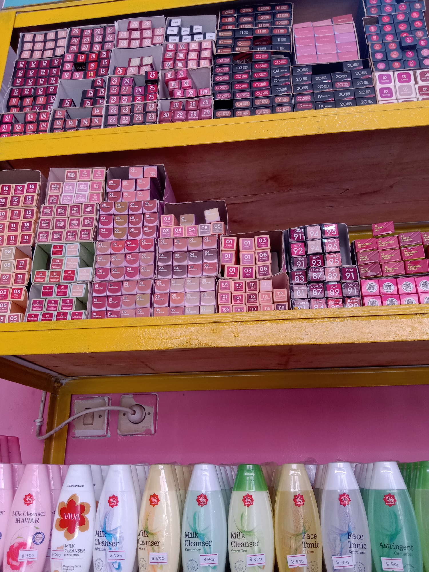 The image shows a retail shelf displaying a variety of cosmetic products, primarily lipsticks and facial cleansers. The products are neatly arranged on two levels of shelving. The top shelf displays rows of boxed lipsticks with numerical and color indicators on their packaging. Brands like "Viny," "Red," "Brown Sugar," "Pink Lava," and "Forest Berry" are visible. Some lipsticks have numerical labels such as "12," "19," "20," "22," "23," "24," "25," "26," "28," "29," "30," "01," "02," "03," "04," "05," "06," "07," "08," "09," "10," "11," and "15." Further to the right, other boxed lipsticks are visible with numerical identifiers from "01" to "06" and "09" to "11" in various shades. Another section shows lipsticks labeled "91," "92," "93," "94," "95," "83," "87," "89," "91," "81," and "82." Brands like "Opal," "Azotic," "Beryl," "Amber," "Pirus," "Zamrud," "Jade," and "Diamond" are also present. The lower shelf displays rows of VIVA brand facial cleansing products, including "Milk Cleanser" in variants like "Mawar," "Bengkuang," "Spirulina," and "Green Tea," as well as "Face Tonic" in "Lemon" and "Spirulina," and "Astringent Cucumber." Each of these products has a price tag of "8.500" displayed on it. The background of the retail space is a pink wall with electrical outlets and wiring visible. The overall environment suggests an indoor retail setting, possibly a small store or a stall. The location is identified as Sugihwaras, Indonesia.