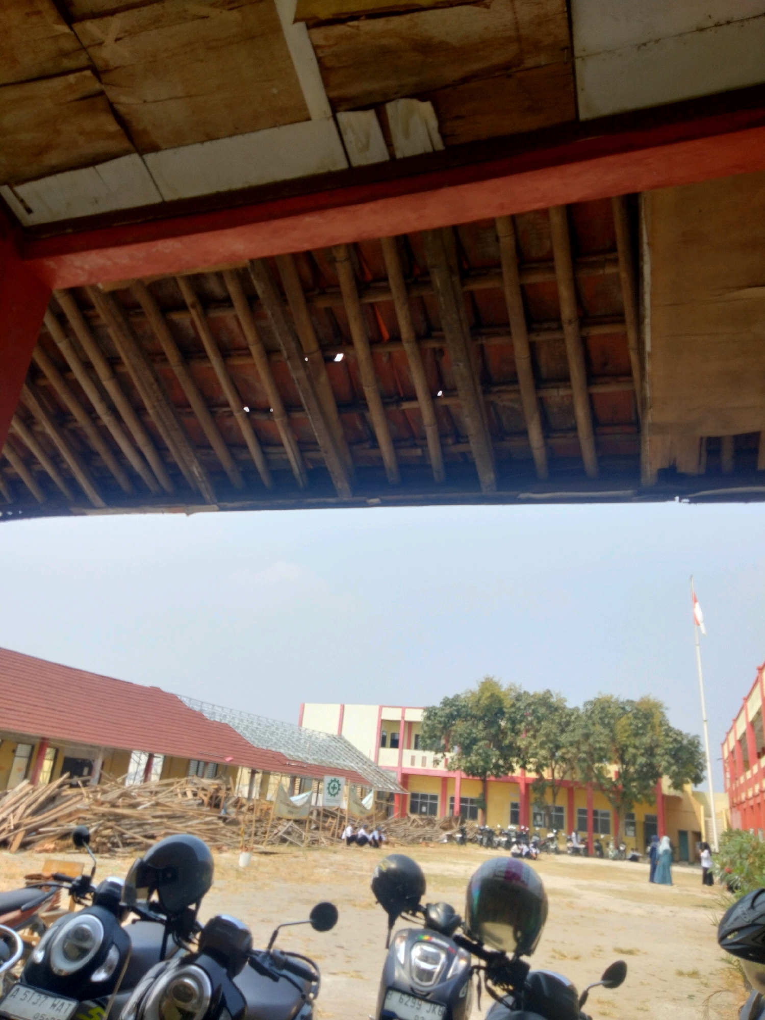 The image depicts an outdoor scene with a building under construction or renovation in the background. In the foreground, several motorcycles are parked. The building appears to be a school or educational institution, with students visible near the entrance. A flag pole with a flag is also present. The overall environment suggests a daytime setting in a somewhat rural or suburban area, possibly in Suka Sari, Indonesia, given the context provided. The construction materials, such as wooden planks, are scattered around the building. The sky is clear and bright.
