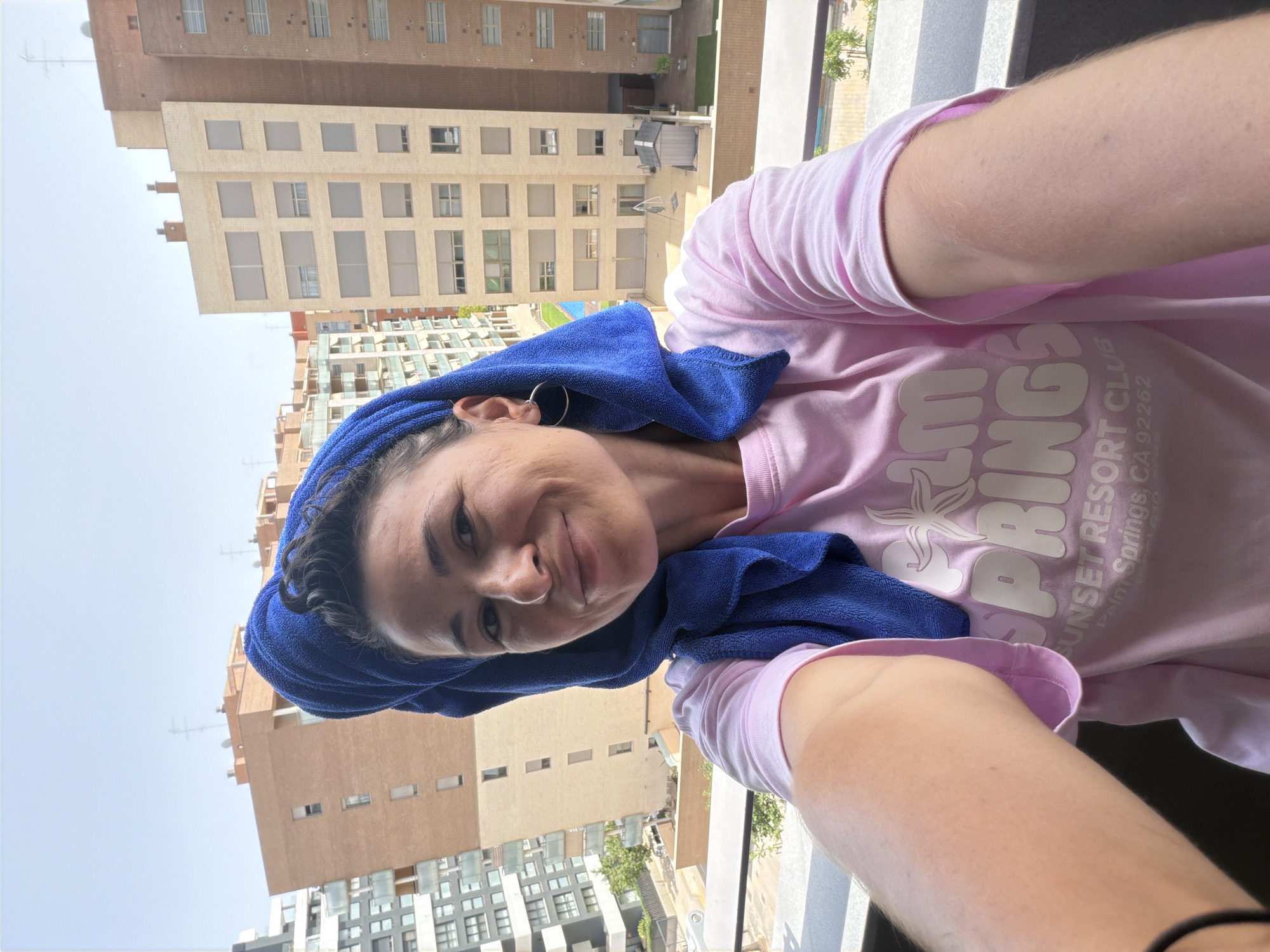 In the foreground, a woman with dark hair wrapped in a blue towel and wearing a pink t-shirt with "Palm Springs Sunset Resort Club" printed on it takes a selfie. She is standing on a balcony with a railing. In the background, there are several apartment buildings of varying heights and architectural styles, set against a clear sky. The scene appears to be in an urban or suburban environment.