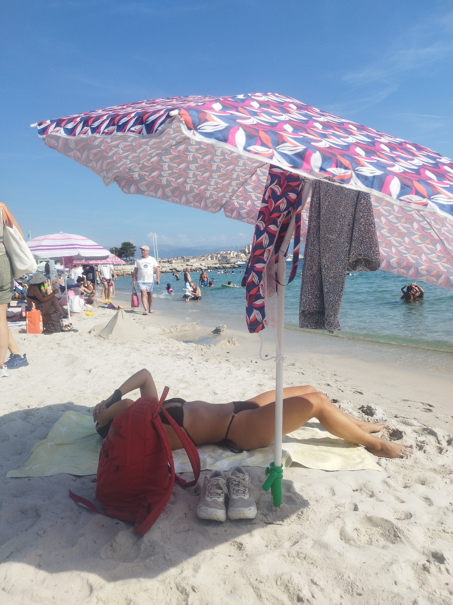 The image captures a sunny day at a beach in Antibes, France. In the foreground, a woman is lying on a yellow towel on the sand, wearing a bikini. Her head is turned away from the camera, resting on her hands. A red backpack and a pair of sneakers are placed next to her. An open beach umbrella with a colorful pattern of red, blue, and white leaves shades her. Two pieces of clothing are draped over the umbrella pole. The sand is light-colored and textured with footprints and shadows. In the mid-ground, other beachgoers are visible. Some are sitting on the sand under another umbrella, while others are walking towards or are in the shallow, clear blue water. A man in a white t-shirt and shorts is walking along the beach, carrying a pink bag. In the background, there is a coastline with buildings and hills under a bright blue sky. The water is calm, and a few boats are docked in the distance.