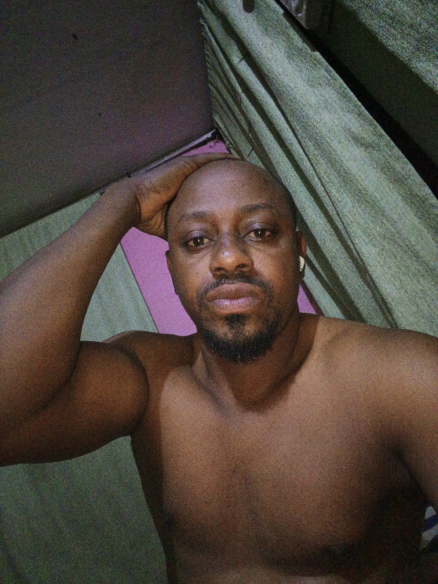 A close-up, low-angle portrait captures a bare-chested man of African descent, looking directly at the camera. He appears to be in his 30s or 40s, with a bald head and a short, dark goatee. A white earbud is visible in his right ear. His left arm is bent, with his hand resting gently on the side of his head. The man's expression is neutral and direct, suggesting a posed moment or a selfie.
The background features draped green fabric, likely curtains, on the right side and behind his right shoulder. To his left and behind his arm, a vibrant pink surface is visible, contrasting with the green. The upper left portion of the image is darker and indistinct. The lighting is somewhat dim, resulting in a slightly grainy image quality, indicative of an indoor setting, possibly during the evening or night. No text is visible in the image. The scene is set in Gwagwarwa, Nigeria.