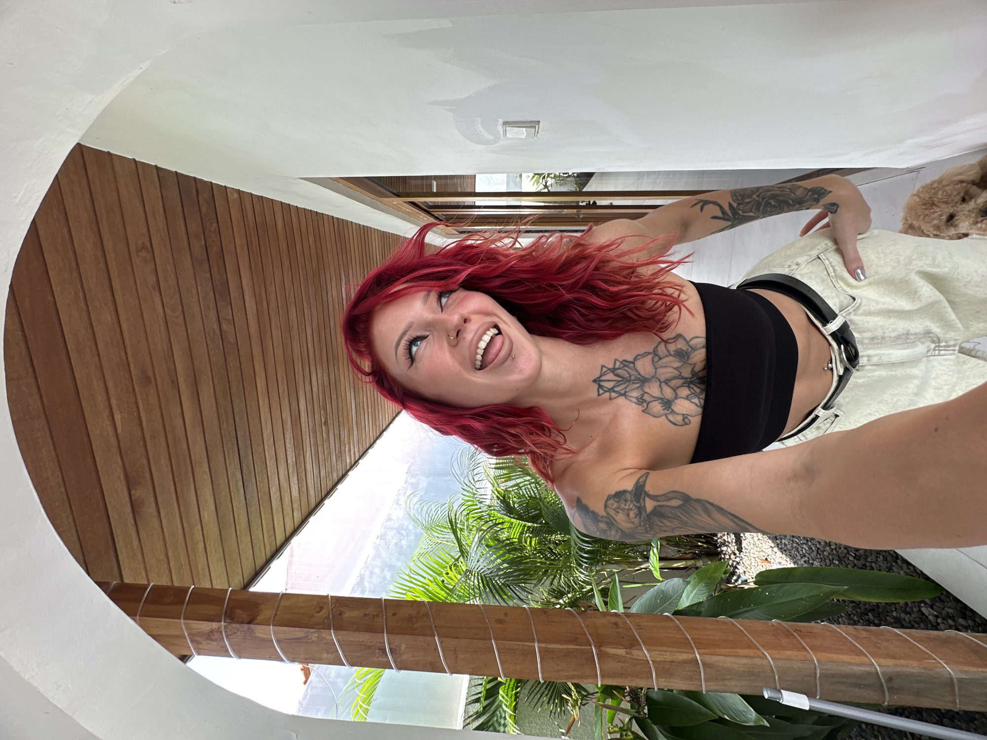 A selfie-style, eye-level photograph depicts a light-skinned person with vivid red hair and tattoos on their arms and chest. They are sticking their tongue out and smiling. The person is wearing a black tube top and high-waisted white pants with a black belt.
The scene appears to be outdoors, framed by an architectural archway. The archway is composed of white plaster and wood paneling. Beyond the archway, greenery and tropical vegetation are visible. Part of a small dog is visible behind the person's pants.
The location is indicated as North Kuta, Indonesia, inferred from the tropical flora visible.