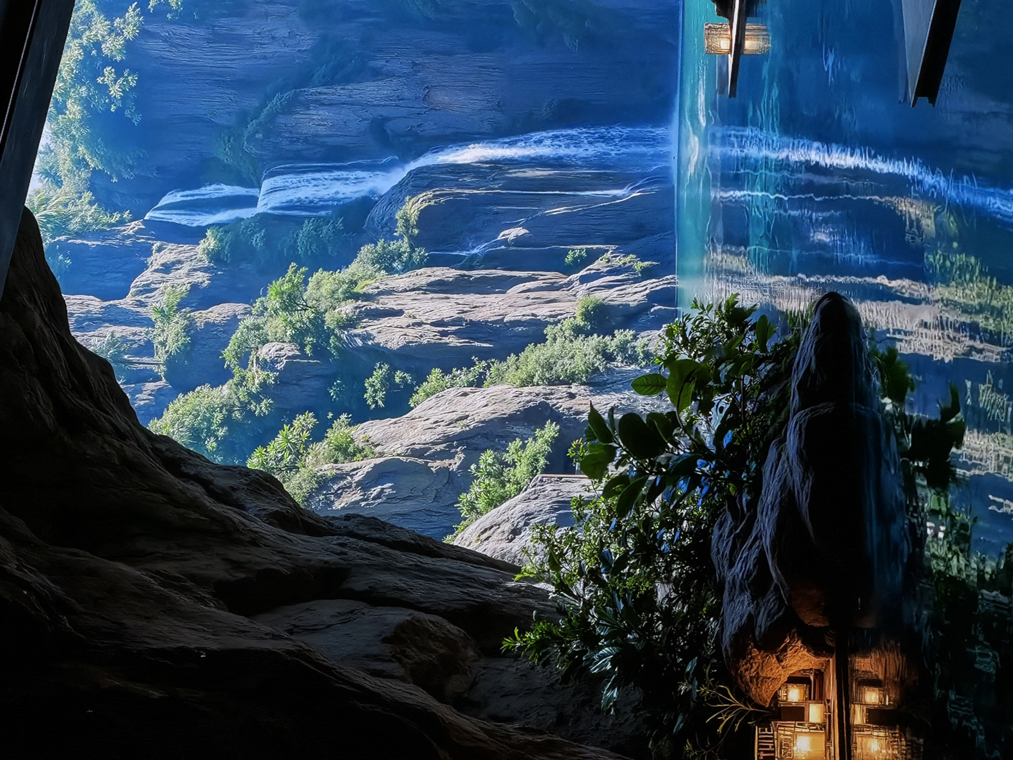 This image captures a dramatic and immersive scene, likely part of a themed attraction or exhibit in Sofiivska Borshchahivka, Ukraine. The primary subject is a dynamic blend of natural-looking landscapes and innovative artificial elements.
On the left and central parts of the frame, a multi-tiered canyon or gorge is depicted, characterized by imposing rocky cliffs and ledges in shades of grey and brown, partially covered with vibrant green foliage, including bushes and small trees. Several waterfalls cascade down the rock faces, appearing as white, foamy streaks against the blue-tinged water, suggesting significant movement and volume. The lighting on these natural elements suggests a bright daytime, with sunlight highlighting the textures of the rocks.
The right side of the image features a striking artificial element: a tall, vertical, luminous blue screen or projection surface displaying shimmering, swirling patterns that mimic flowing water, ripples, and reflections. This creates the illusion of a monumental waterfall or a dynamic reflective pool within an indoor setting. In the foreground on the right, a large, dark, sculpted rock formation, possibly artificial, is adorned with green foliage and partially obscures the blue screen. Integrated into the base of this formation, several intricately designed, warm glowing lanterns are visible, casting a soft light. Faint, vertically stacked white text "T H E" is discernible on a dark element near the bottom-most lantern. The foreground on the left is a dark, shadowy area, revealing rough, textured rock, possibly a viewing platform or a cave entrance.
The scene creates a sense of wonder and immersion, leveraging both realistic landscape design and advanced projection technology to transport the viewer to a grand, natural-like environment. The juxtaposition of natural light and shadow with artificial illumination and effects suggests a carefully curated visual experience. No people are visible in the frame, allowing the focus to remain on the scale and detail of the environment.