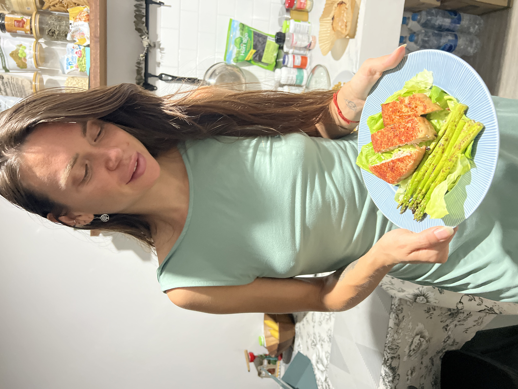 A young woman with long, dark brown hair, wearing a light green sleeveless top or dress, is depicted holding a plate of food. Her eyes are closed, and she has a subtle, content smile on her face, suggesting a moment of peace or satisfaction. She is holding a light blue plate adorned with thin white lines, featuring a healthy meal. The meal consists of three rectangular pieces of pan-fried protein, possibly salmon or tofu, lightly seasoned with a reddish spice. These are served on a bed of fresh green lettuce alongside several stalks of cooked green asparagus.
The background reveals an indoor kitchen or dining area. To the left, shelves are visible, holding clear glass jars filled with various dried foods, grains, and spices. A green plastic bag with the brand "Herbalife" is prominently displayed on a shelf. Several bottles and containers, likely supplements or spices, are arranged on a white tiled wall. A black frying pan rests on a countertop. In the deeper background, stacks of plastic water bottles can be seen. Part of a table or counter covered with a grey and white floral patterned fabric is visible in the lower right. The overall impression is a domestic setting, likely in บ้านบางรักษ์, Thailand, where the woman is presenting or about to enjoy a freshly prepared meal.