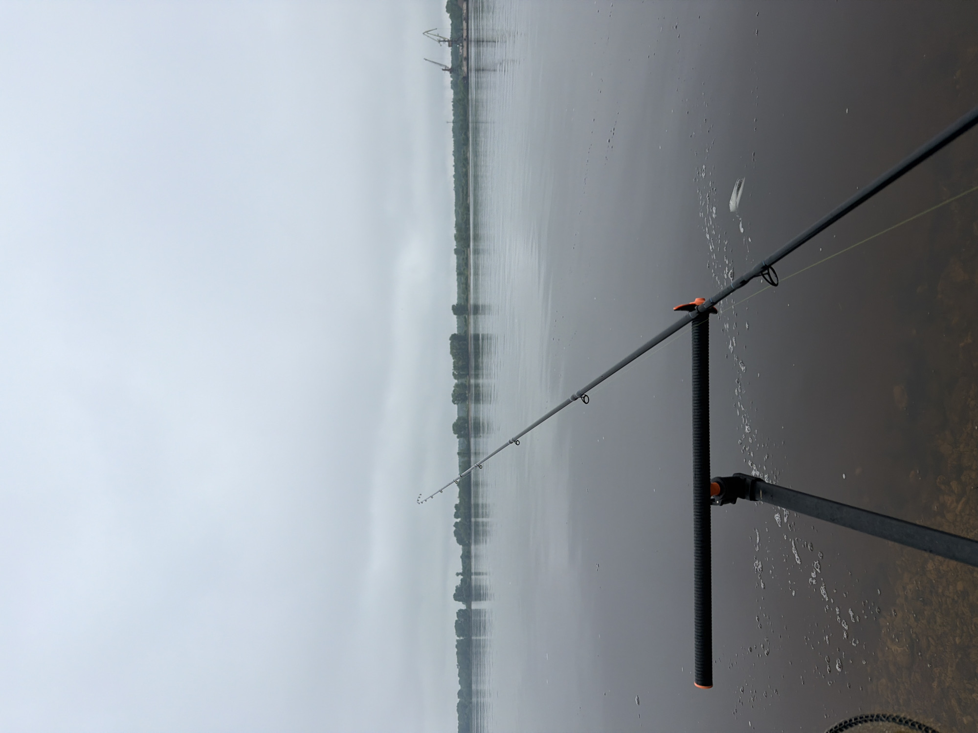 The image depicts a serene fishing scene by a wide body of water under a heavily overcast sky. In the foreground, a black fishing rod is set up, supported by a dark rod rest. The rod features an orange-tipped handle and orange accents near the reel seat, and its tip extends over the water. A thin, light-colored fishing line is visible, exiting the rod and disappearing into the calm, grey water.
The water surface shows subtle ripples and scattered patches of white foam or bubbles, suggesting slight movement or a recent disturbance. The midground is dominated by the expansive water, which reflects the dull, uniform light of the sky and the distant shoreline.
Across the horizon, a low-lying, dark green treeline marks the far bank of the water body. Above the trees, on the distant shore, two large industrial cranes are visible, hinting at a port or industrial activity in the area. The sky is a uniform, pale grey, completely covered in clouds, creating a subdued and quiet atmosphere.
There are no people visible in the image. The scene conveys a sense of calm and patience, typical of fishing. The time of day appears to be daytime, but the heavy cloud cover prevents direct sunlight. No text is visible. The setting is consistent with a large river or lake, such as found in Chaikovsky, Russia.