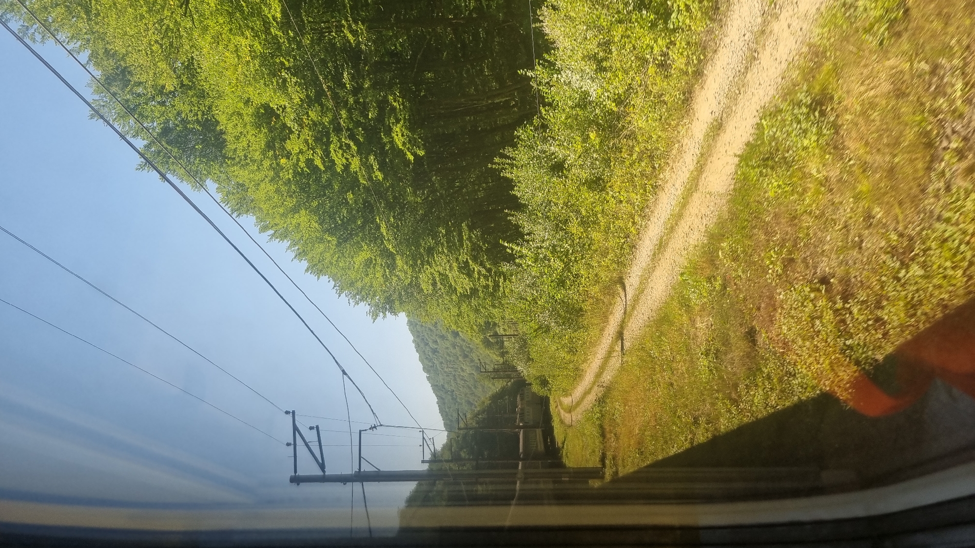 This is a vertical, eye-level, full-shot photograph taken from a moving train, looking out its window. The scene depicts a sun-drenched rural landscape in the vicinity of Han'kovytsya, Ukraine. The foreground is dominated by the train window frame on the left, reflecting the bright sky and suggesting motion. A dirt road curves through lush green grass and foliage, leading into a dense forest that covers rolling hills in the background. Overhead, power lines stretch across the clear blue sky, supported by poles that are partially visible. The natural light suggests it is daytime, likely late morning or early afternoon. There are no people visible in the image.