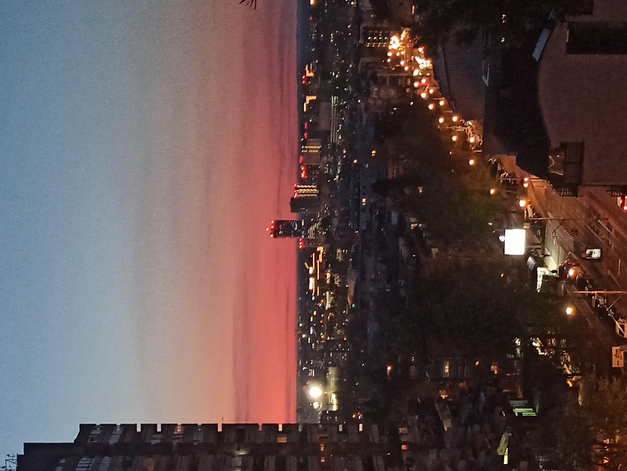 The image is a rotated, high-angle view of Vozdovac Urban Municipality, Serbia at dusk. The sky is a gradient, transitioning from blue to reddish-orange above the city. A cluster of high-rise buildings stands in the middle ground, illuminated by city lights. A major road runs along the right edge of the image, lined with streetlights and active vehicles. Buildings are situated at the top and bottom of the frame.