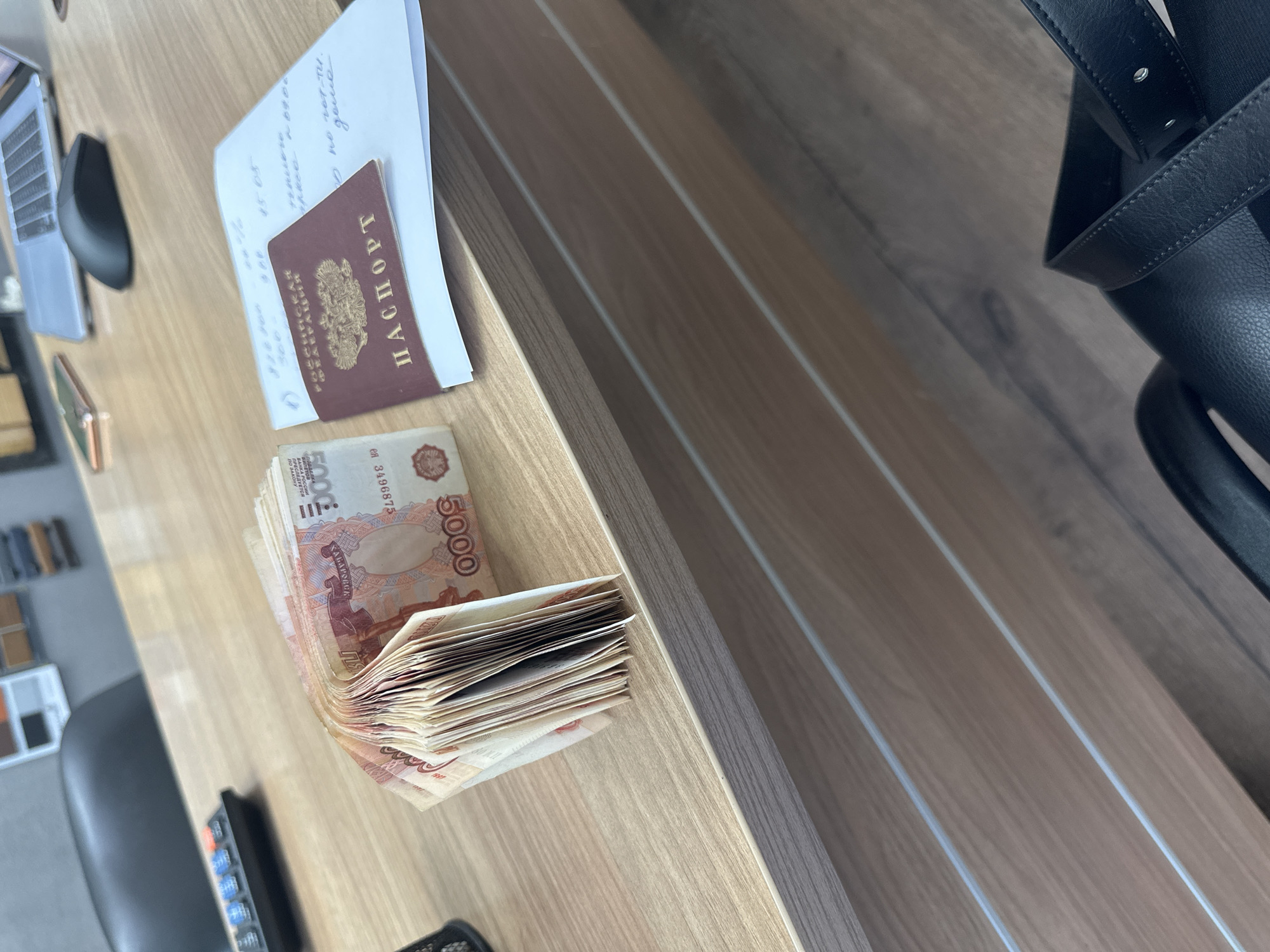 The image depicts a desk or table with a collection of items. Located in an unidentified location in Russia, but given the passport, possibly the city of Izhevsk, a Russian passport lies on top of a piece of paper with handwritten notes. Next to the passport is a stack of Russian currency, specifically 5000 ruble banknotes. A laptop and a mouse are visible in the background. A black chair is also partly visible. The objects are arranged on a wooden surface, presumably the desk or table.