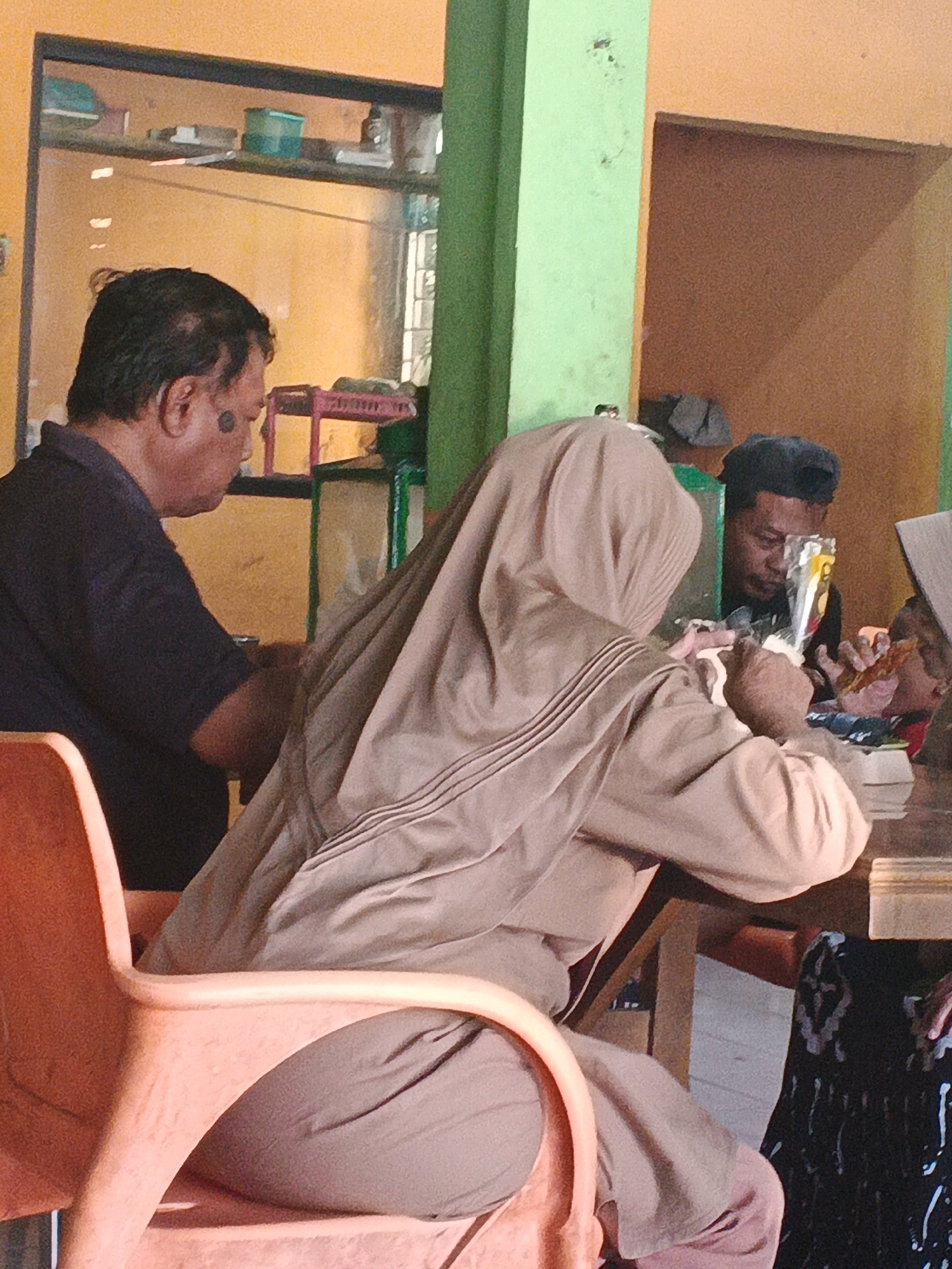 A medium shot captures a group of individuals at a restaurant in Ungaran, Indonesia. In the foreground, an orange plastic chair is visible, partially occupied by a person wearing a light brown hijab and matching attire, their hands are engaged with food. To their left, a man with dark hair and a dark shirt is seated, his profile visible as he looks down. Behind them, another man wearing a dark cap and black shirt is also seated, looking towards the table. The environment appears to be an open-air or semi-open-air eatery with orange and green painted walls, and a glass display cabinet in the background. The lighting suggests an indoor setting during the day.