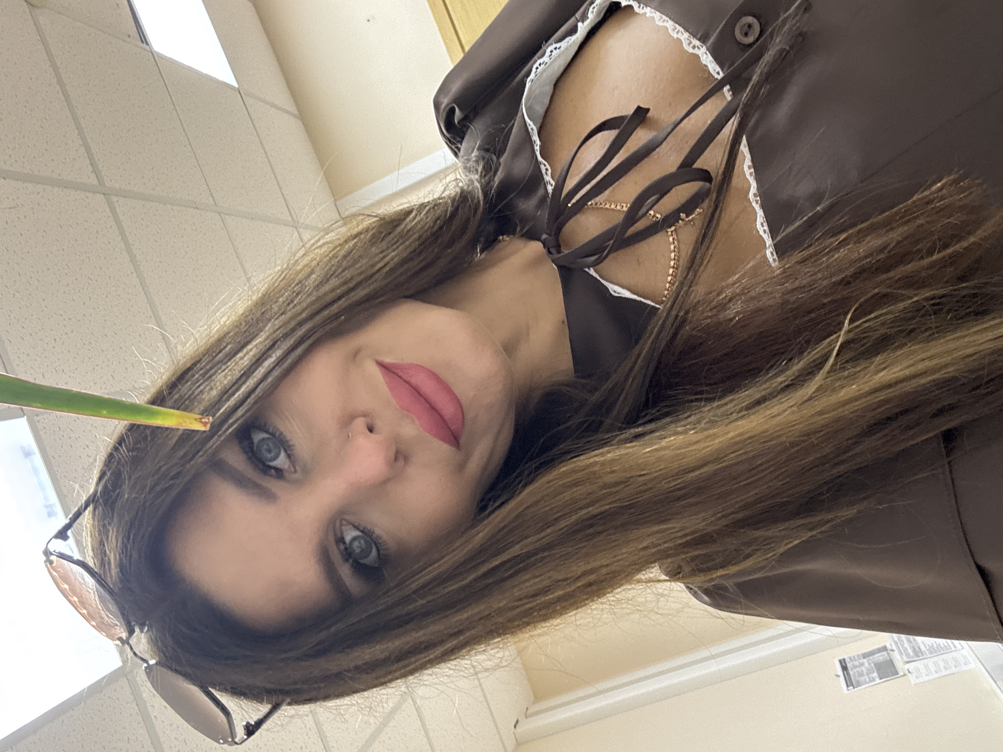 A woman with long brown hair and blue eyes, wearing a brown dress with white lace trim and a gold chain necklace, poses for a selfie. She is wearing sunglasses on her head, and a green leaf is held near her face. The background appears to be an indoor setting with a tiled ceiling and beige walls. The image does not provide any information about the location in Chaikovsky, Russia.
