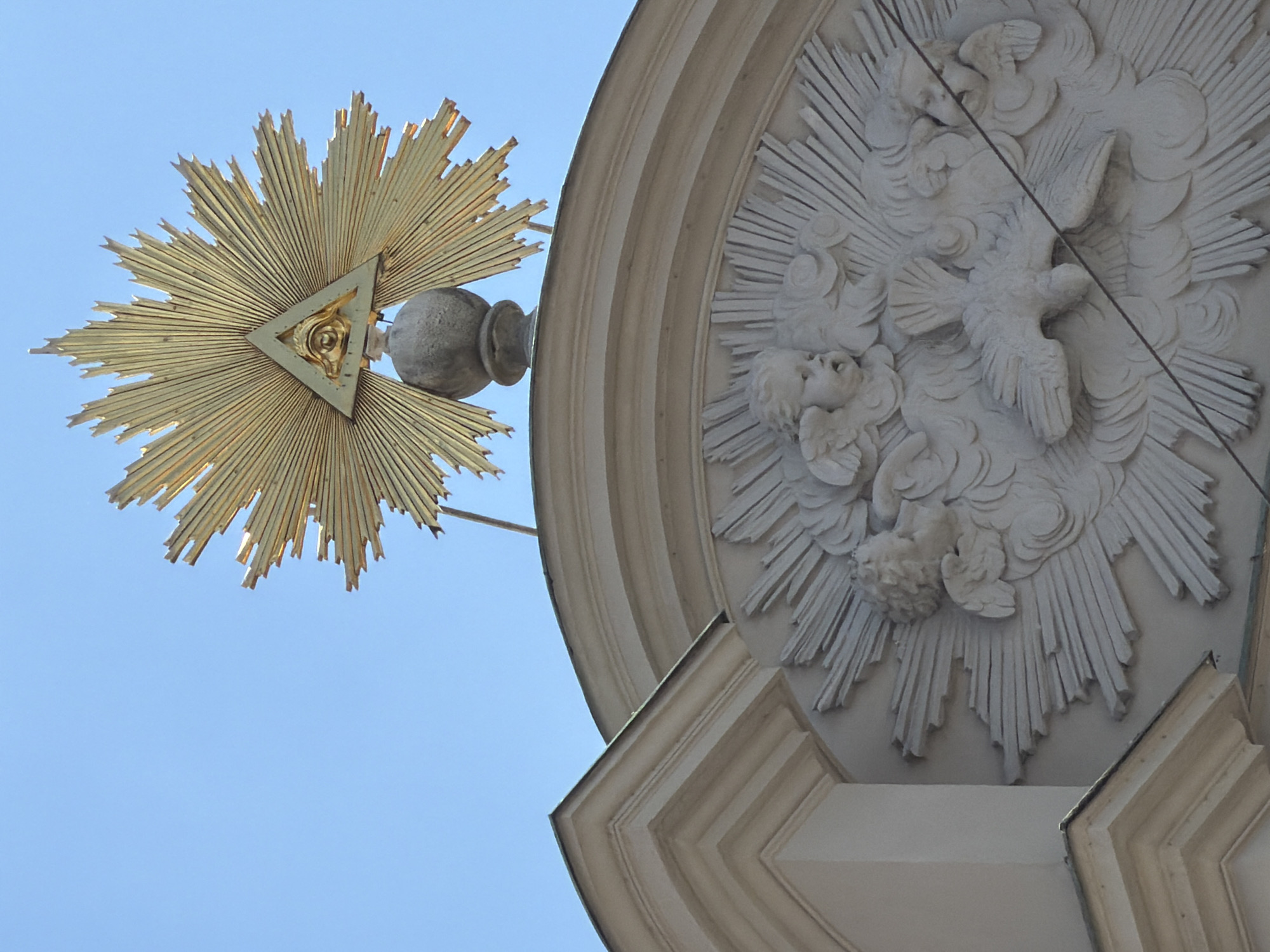 The image shows the top portion of a building facade in Munich, Germany, under a clear blue sky. Dominating the upper part of the image is a golden, sunburst-like emblem with rays emanating outwards. At the center of the sunburst is a triangle containing an eye, a symbol often associated with divine providence. Below this emblem, a sculpted relief adorns the facade. This relief depicts several cherubic figures and a dove, all surrounded by radiating lines and cloudy forms. The architectural style suggests a Baroque or Rococo influence. The building appears to be an older, ornate structure, likely a church or a historic civic building, given the religious symbolism. The lighting indicates it is daytime, with the sun casting a warm glow on the golden emblem.