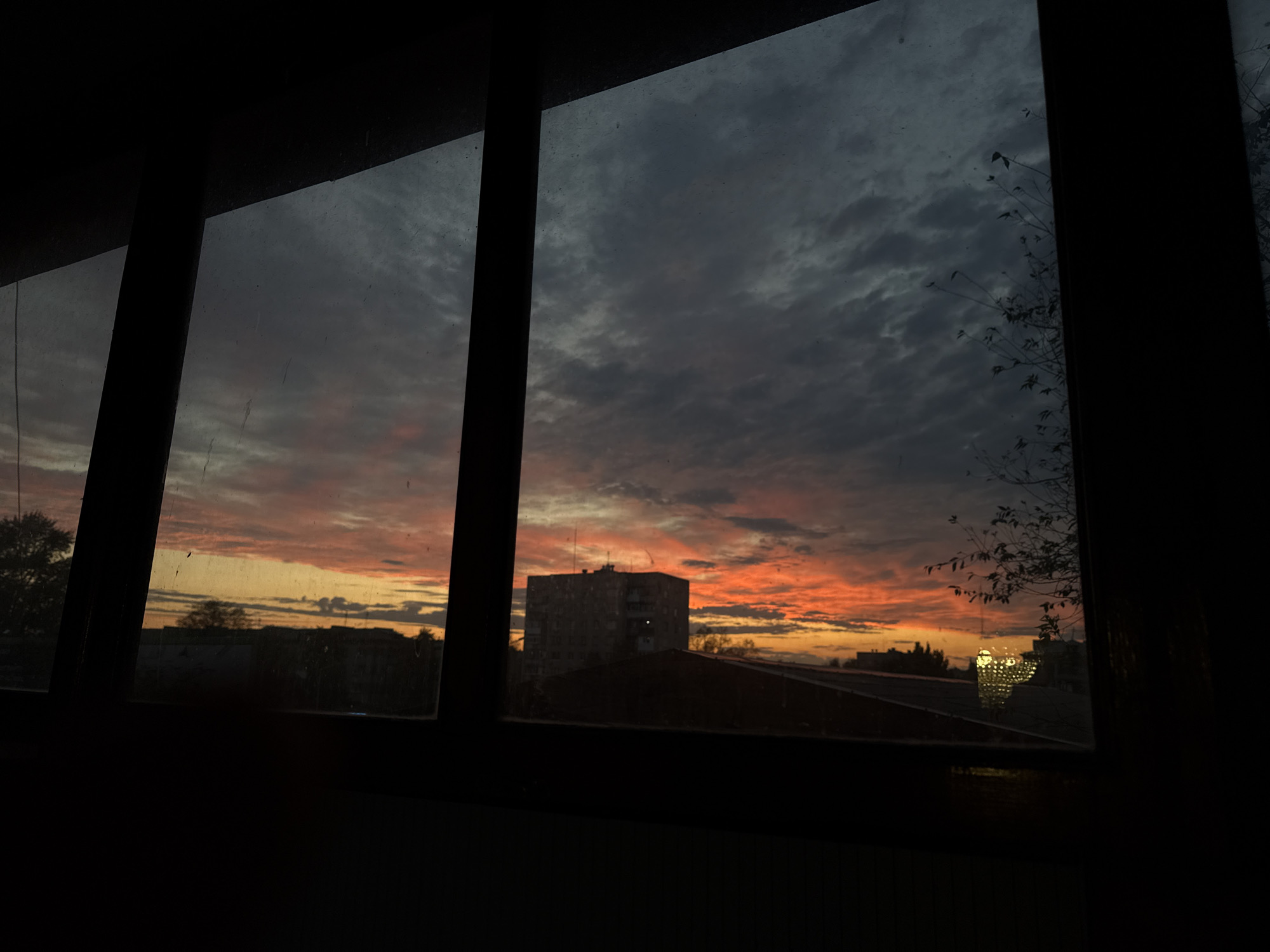 The image shows a view through a window of a sunset over the city of Luhansk, Ukraine. The sky is filled with dramatic clouds colored in shades of orange, red, and gray, indicating a vivid sunset. In the foreground, the dark silhouettes of window frames divide the view into several panes. Through the window, various buildings can be seen, including a multi-story apartment building and other structures that appear to be residential or commercial. Trees and branches are also visible, some silhouetted against the bright sky. A bright, glowing light reflects in the window on the right side of the image, possibly from a streetlamp or building lights. The overall scene is one of twilight transitioning into evening, with the urban landscape partially obscured by the darkening sky and the window's perspective.