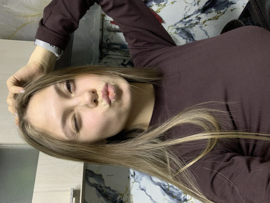 The image displays a young adult female subject, positioned centrally and captured from the chest up, in an indoor setting. She has long, straight, light brown to blonde ombre hair, parted on her left, draped over her shoulders. Her right eye (viewer's left) is closed in a wink, while her left eye (viewer's right) is open. Her lips are pursed into a pouting shape, revealing a small silver labret piercing on the lower lip and a subtle metallic septum piercing. Her left hand (viewer's right), adorned with light-colored fingernails, is raised to her head, with fingers resting on the top of her hairline. She is wearing a dark maroon, long-sleeved, crew-neck top. A light grey wristband or watch strap is visible on her right wrist (viewer's left). The background on the upper left shows a portion of a light-colored kitchen-style cabinet with a metallic handle. To the right and behind the subject, a wall or backsplash is visible, featuring a dark grey and white marbled pattern with faint light brown or gold veining. The precise location within the city of Fokino, Russia, cannot be determined from the visual information; however, the environment is consistent with a residential kitchen or a room equipped with kitchen-like cabinetry and finishes.