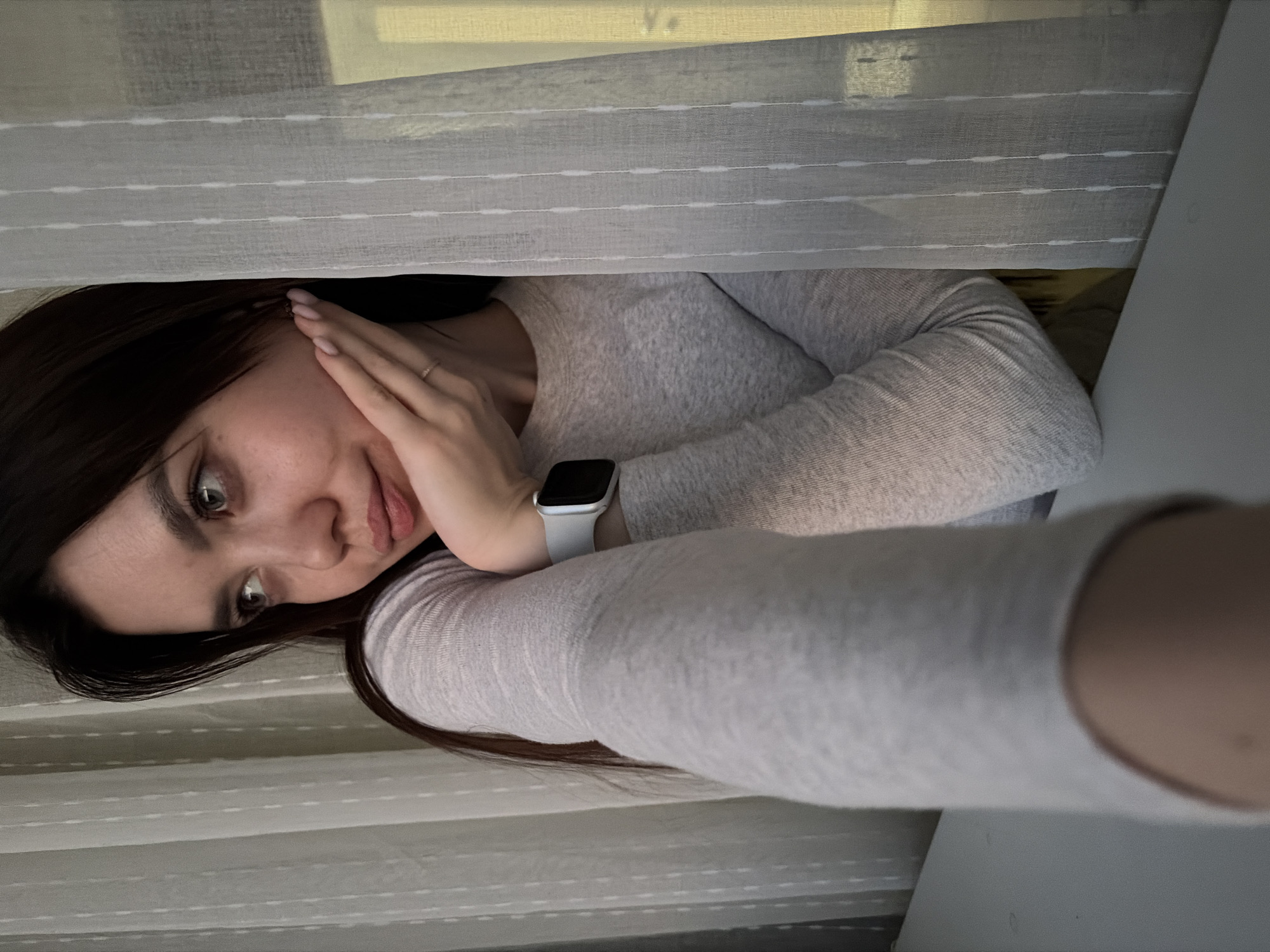 A vertically oriented selfie captures a young woman indoors in Chaikovsky, Russia. She has long, dark hair, fair skin, and light-colored eyes, looking directly into the camera with a soft, slightly pensive expression. Her right hand is gently placed near her face, revealing manicured nails with a light pink polish and a thin ring on her finger. She wears a light grey, long-sleeved top and a light-colored (possibly white or light grey) smartwatch on her left wrist. The woman appears to be in a relaxed, possibly reclining position. The background consists of light-colored, sheer curtains or blinds with horizontal striped patterns, indicating a window and an indoor setting. A solid light-colored surface, possibly a table or wall, is visible on the right side of the frame. The lighting is soft and diffused, suggesting it is daytime. There is no visible text in the image.