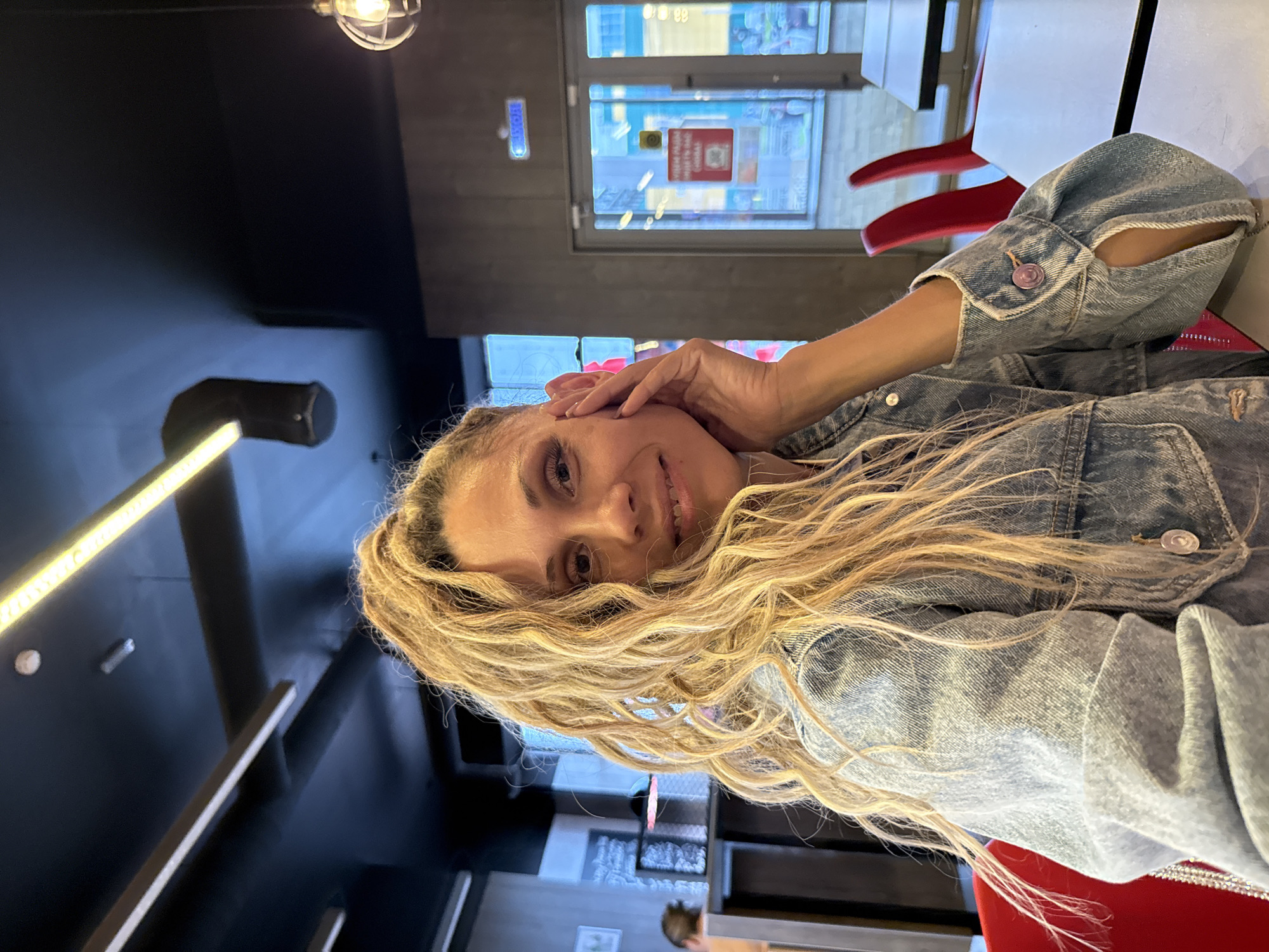 A medium-shot, head-and-shoulders, indoor selfie shows a woman with blonde, curly hair tied in a high ponytail, wearing a denim jacket. She is smiling and has her right hand on her face, near her ear. The background appears to be a restaurant with dark ceilings, industrial lighting, and red chairs. A door with signs in Russian is visible behind her. The location is Ignatovo, Russia.