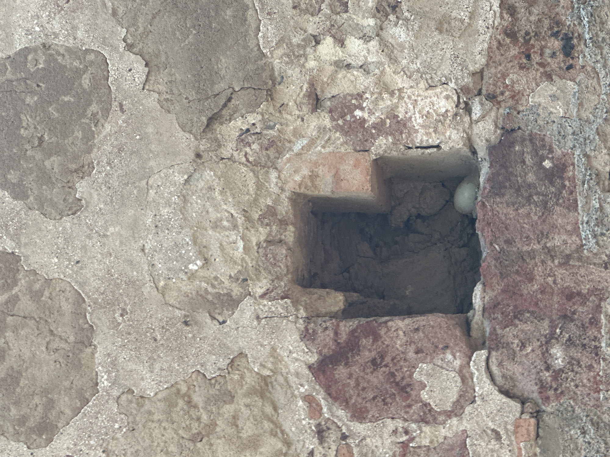 A close-up shot reveals a section of an aged, weathered brick wall. The wall is constructed with a combination of irregularly shaped stones and fragmented bricks, exhibiting significant erosion and deterioration of the mortar and surface layers. A square cavity is visible within the wall, possibly a missing brick or intentional opening, exposing the interior of the wall and revealing a small, round, white object partially obscured within the recess. The coloration of the wall varies, with some areas exhibiting a reddish-brown hue from the bricks and other parts showing a greyish-white from the deteriorated mortar and stone. The surrounding environment and specific location in Tlalnepantla, Mexico, cannot be determined from the provided image. No people or interactions are visible.