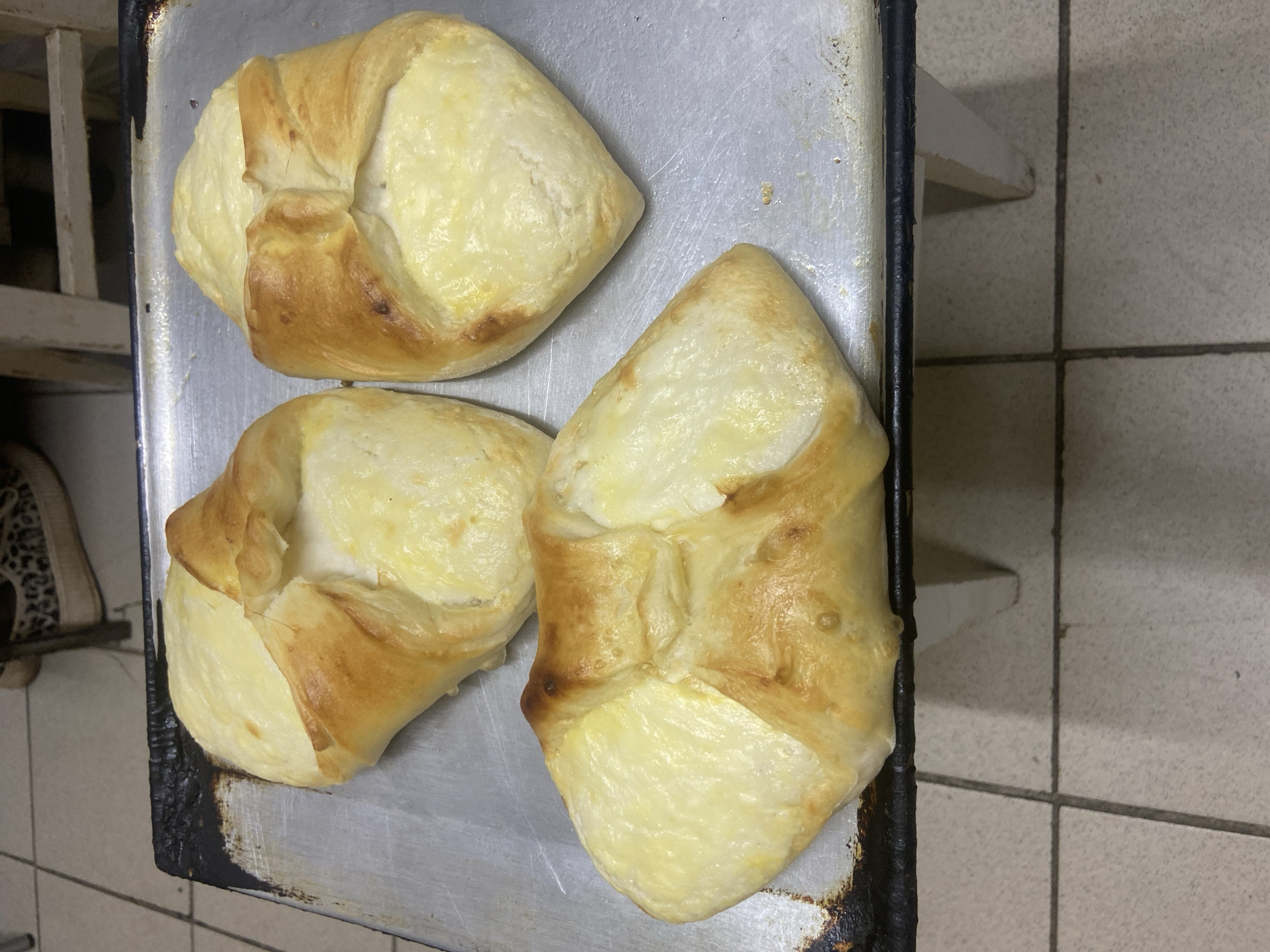 The image shows three freshly baked pastries arranged on a tarnished metal baking tray. The pastries have a golden-brown crust and a creamy filling that appears to be cheese. The tray is sitting on what seems to be a set of metal shelving. The background includes tiled walls and flooring. I cannot confirm the location is Chaikovsky, Russia, without additional information.