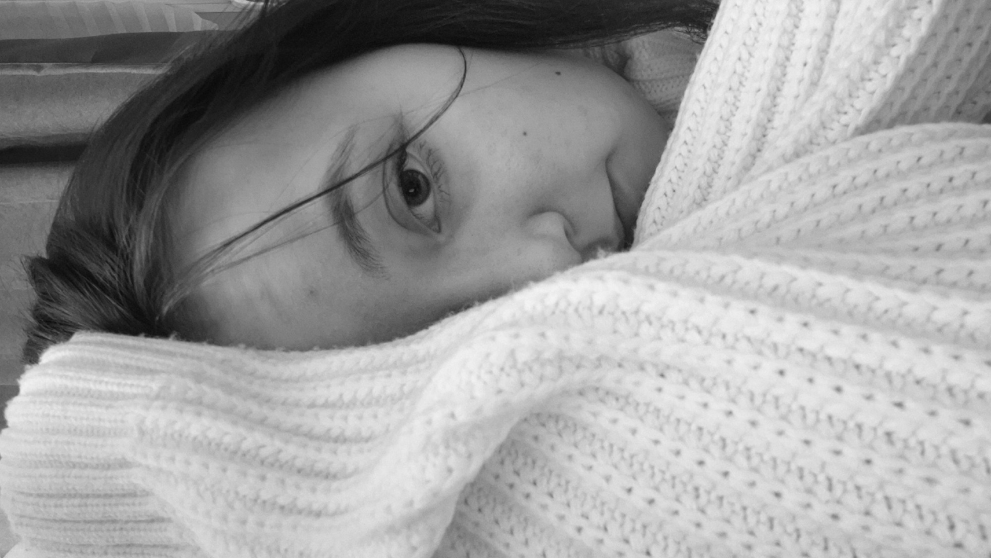 A monochrome, close-up photograph shows a person partially obscured by a textured, knitted garment. The person's face is turned towards the camera, with one eye visible. Strands of dark hair are visible across the face. A portion of the nose and cheek are also visible, partially hidden by the garment. The background is out of focus. The location is unknown but is speculated to be in Chaikovsky, Russia. No actions or interactions are depicted.