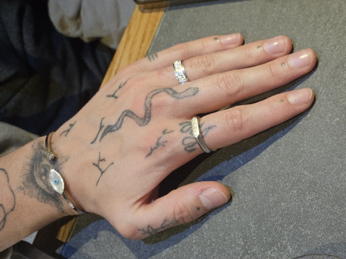 A person's hand rests on a surface, adorned with tattoos and jewelry. A silver bracelet with a blue and white center sits on the wrist, below a sun-like tattoo. A snake tattoo curves across the top of the hand. A ring with inlaid gemstones is on the ring finger, and a silver ring with text etched into it is on the middle finger. The fingernails have small dark spots on them.