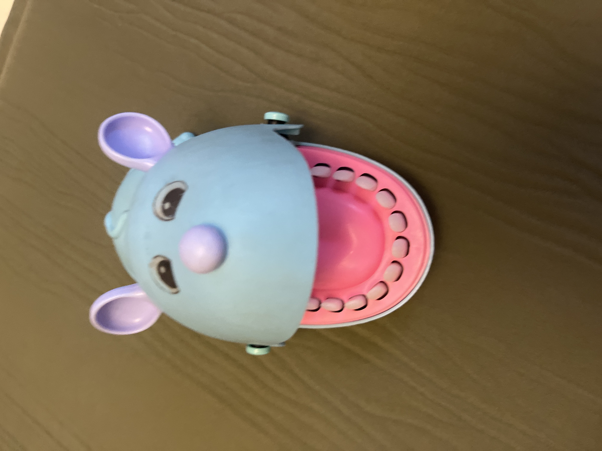 The image shows a close-up, top-down view of a light blue toy with large, round, light purple ears. The toy has two dark eyes and a light purple nose or cheek puff. Its mouth is open, revealing a bright pink interior with a row of white "teeth" along the edge. The toy appears to be made of plastic. The background is a textured, brownish-grey surface, possibly a tabletop or floor. The lighting suggests an indoor environment. There is no visible text in the image. The overall impression is of a stylized animal or creature, possibly a mouse or elephant. The toy is positioned diagonally on the surface.