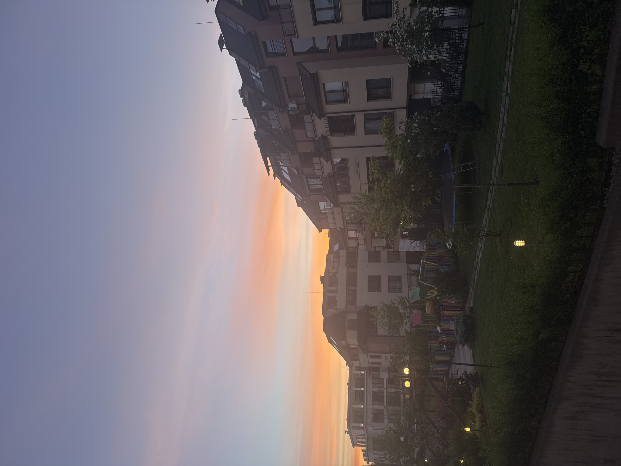 The image shows a row of buildings with a park on the side during sunset in Sofia, Bulgaria. The buildings are multi-story, modern apartment complexes. The sky is a gradient of colors from pale blue to orange, indicating sunset. The park has trees, grass, playground equipment, and lit lampposts. No people are visible.