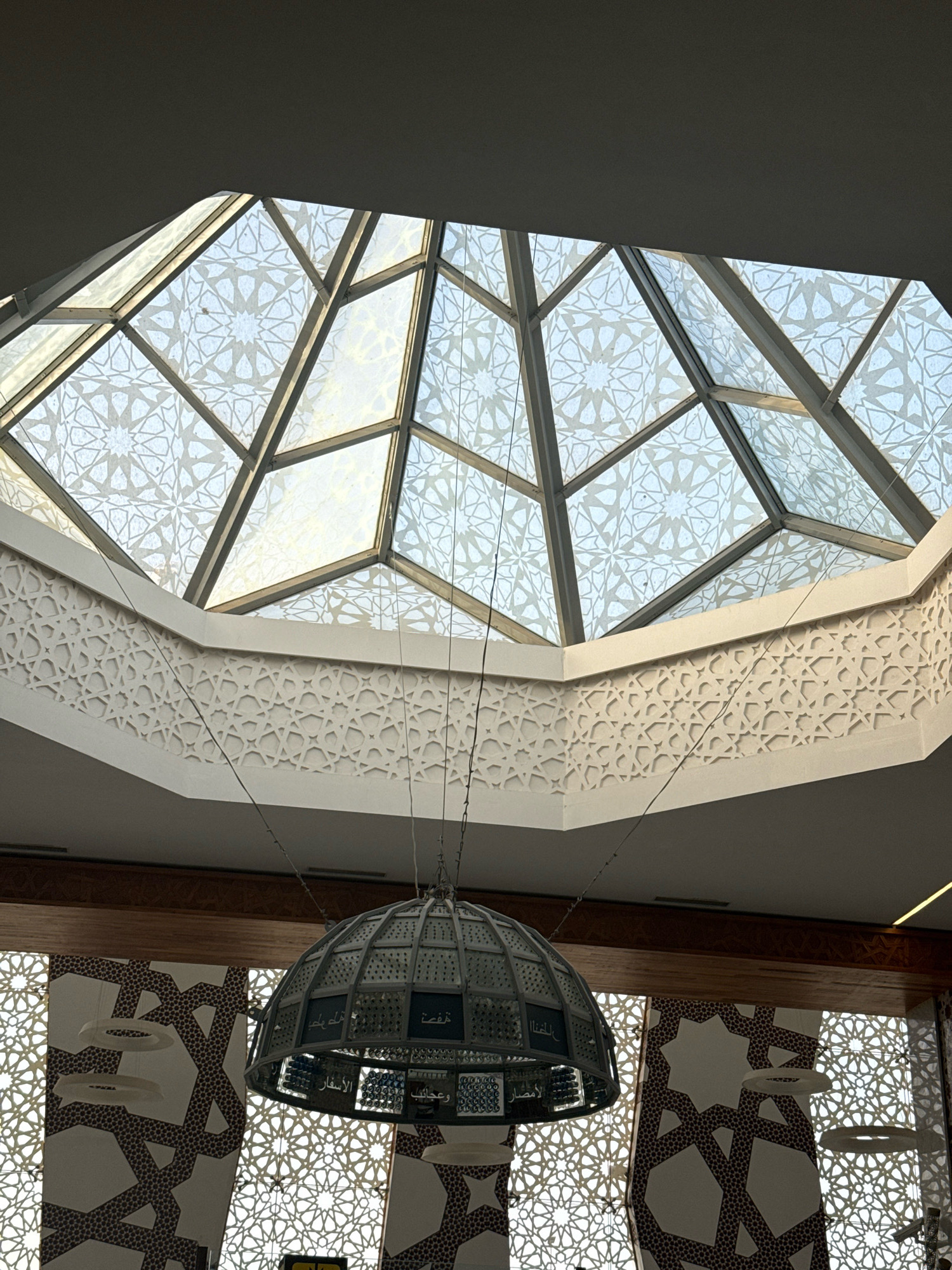 This is a low-angle, interior shot looking up towards a large, geodesic dome skylight made of glass panels, each adorned with a repeating geometric, Islamic-inspired pattern of white on a frosted or semi-transparent background. The metal framework supporting the glass is visible, forming a grid. Below the skylight, a decorative frieze runs along the top of the wall, also featuring a detailed geometric Islamic pattern carved or applied in a raised, white relief.
Suspended from thin wires in the center of the frame is a large, metallic, dome-shaped light fixture. It appears to be constructed from panels, some of which have dark blue inserts with Arabic calligraphy. The light fixture is intricate and multi-faceted.
The background below the frieze shows a section of a wall or partition featuring a large-scale repeating pattern of stylized dark brown or black shapes on a white background. These shapes appear to be interconnected polygons or starbursts, also in an Islamic geometric style.
The overall environment suggests a modern building, possibly a public space like a mosque, cultural center, or a modern architectural space with significant Islamic design influences. The lighting is soft and diffused, coming primarily from the skylight. There are no people visible in the image. The architectural elements suggest a specific location, likely a building within the city of Ouled Tayeb, Morocco, known for its rich Islamic heritage and architectural traditions.