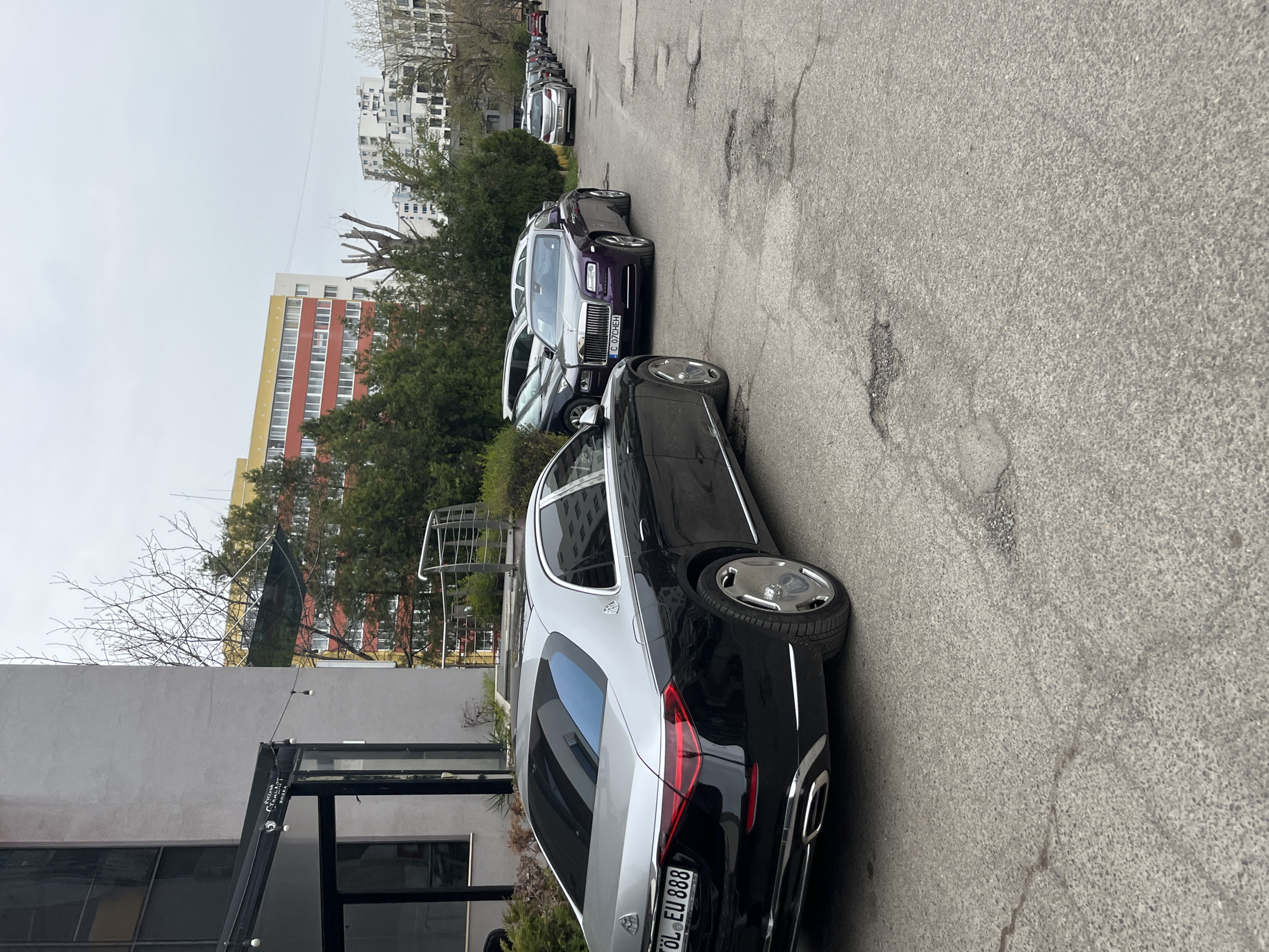 The image shows a street scene in an urban area. The main focus is on a line of parked luxury cars, including a black and silver Maybach and a purple Rolls Royce. The cars are parked on a narrow street with visible pavement damage. In the background, there are buildings, some with visible architectural details such as balconies and windows. Trees are also present, adding a natural element to the urban landscape. The sky is overcast, suggesting a cloudy day. The image is taken at a slightly oblique angle, providing a perspective view of the street. The license plates of the cars are visible, providing specific details about the vehicles. The image is well-lit, with sufficient light to capture details of the cars and the surrounding environment.