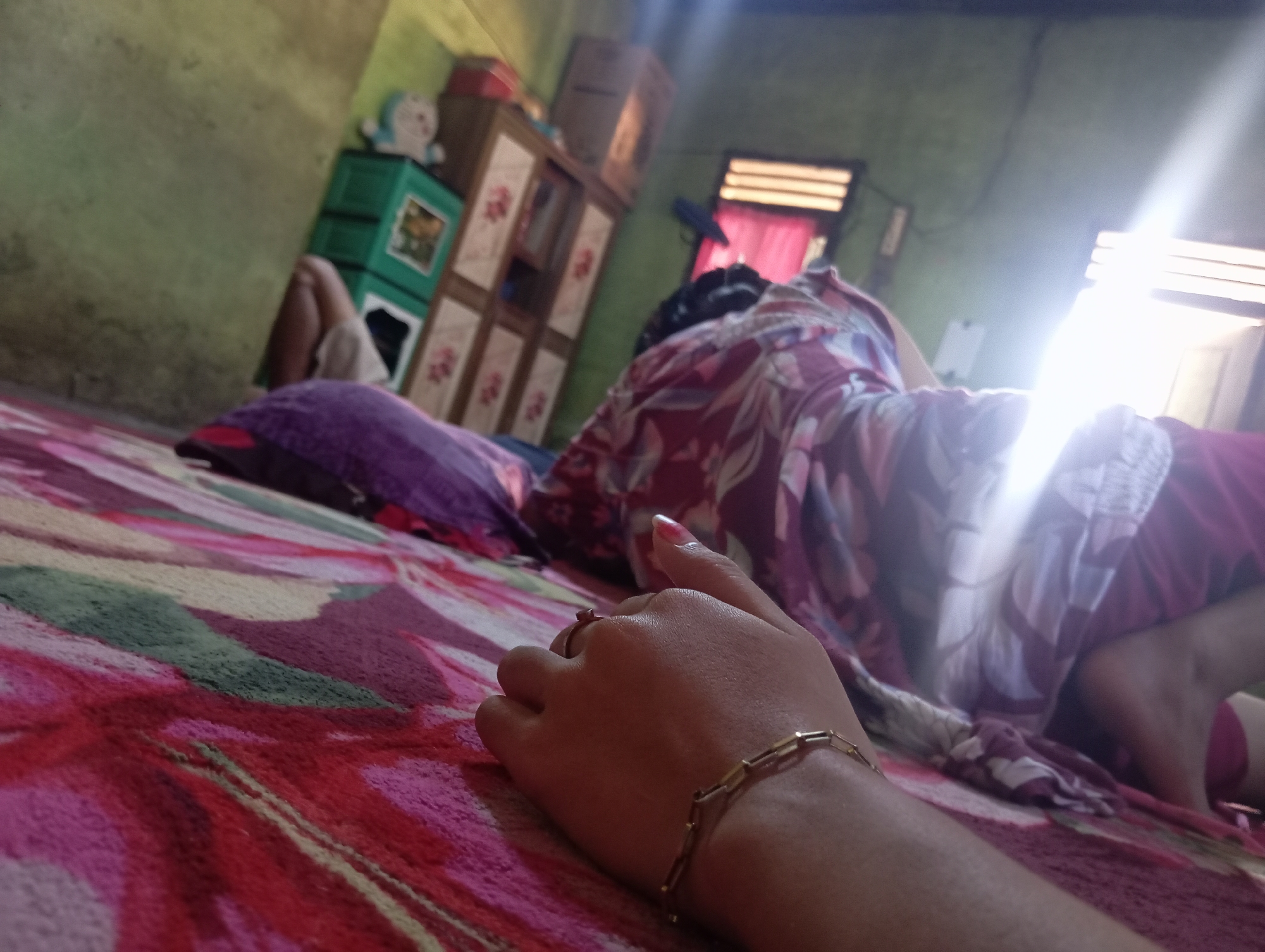 The image depicts an indoor scene in Rejosari, Indonesia. A person is lying on a colorful bed, facing away from the camera. They are wearing a patterned shirt and pink pants. In the foreground, a hand with a ring and a bracelet is resting on the bed. The background shows a green wall with some furniture, including a cabinet with drawers and a toy figure on top. Sunlight is streaming in from a window, creating a bright glare in the upper right portion of the image. The overall impression is of a casual, lived-in space.