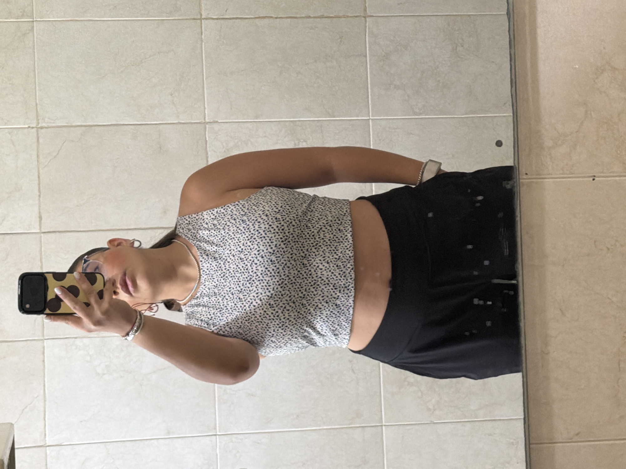 A young person stands in a tiled room, possibly a bathroom, taking a selfie in a mirror. They are wearing a printed crop top and black pants. The phone has a patterned case. The tile pattern in the background appears to be a common style. There is no other information in the image to suggest the location is specifically Puebla, Mexico. The information about Puebla, Mexico is extraneous.