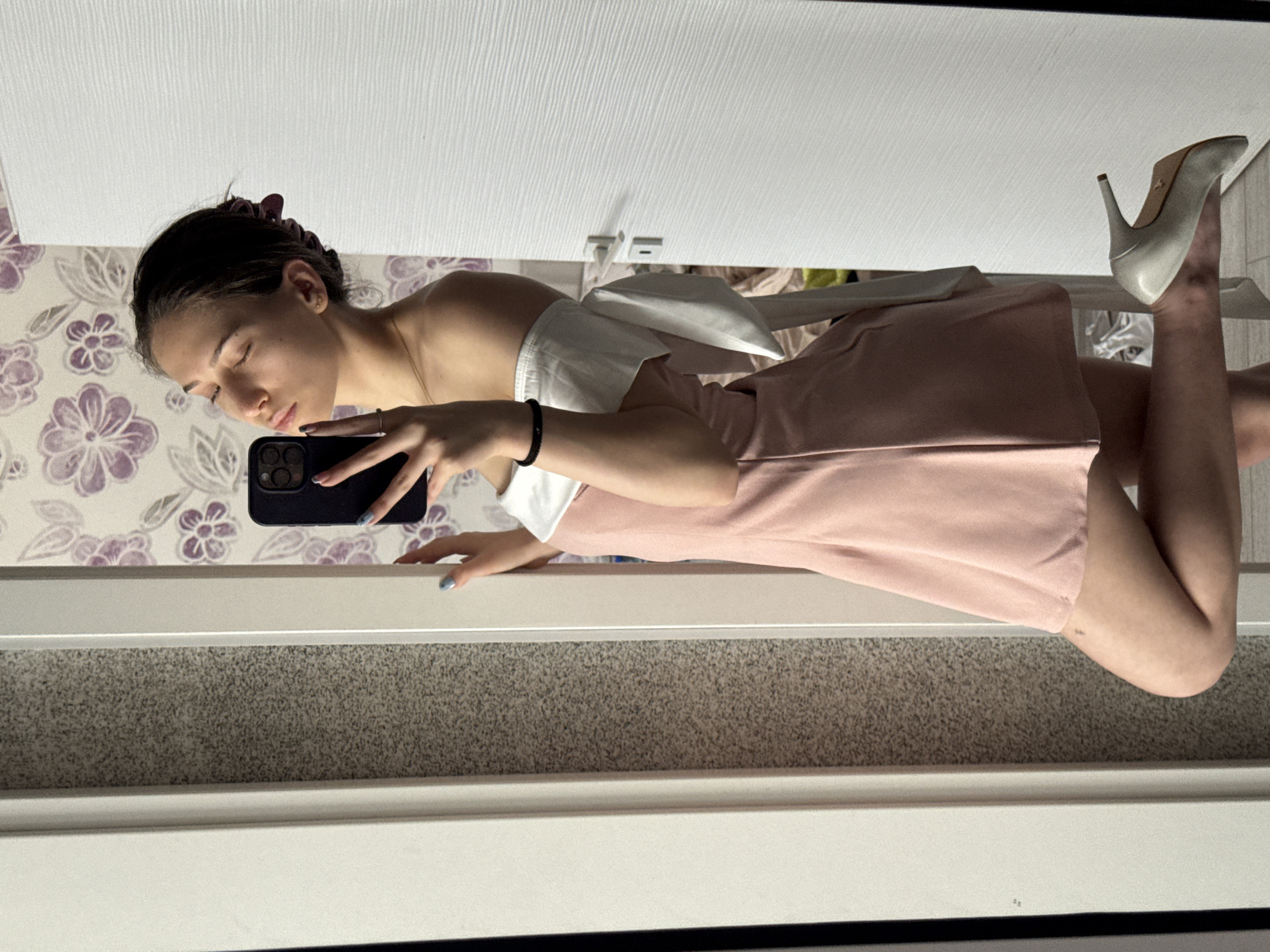 A woman is posing in a mirror. She has dark hair and is wearing a pink dress with white details. She is holding a black smartphone and taking a selfie. She is also wearing white high-heeled shoes. The background includes a white door, patterned wallpaper with flowers, and a gray carpet. There is no information to suggest that this image was taken in Sofia, Bulgaria.
